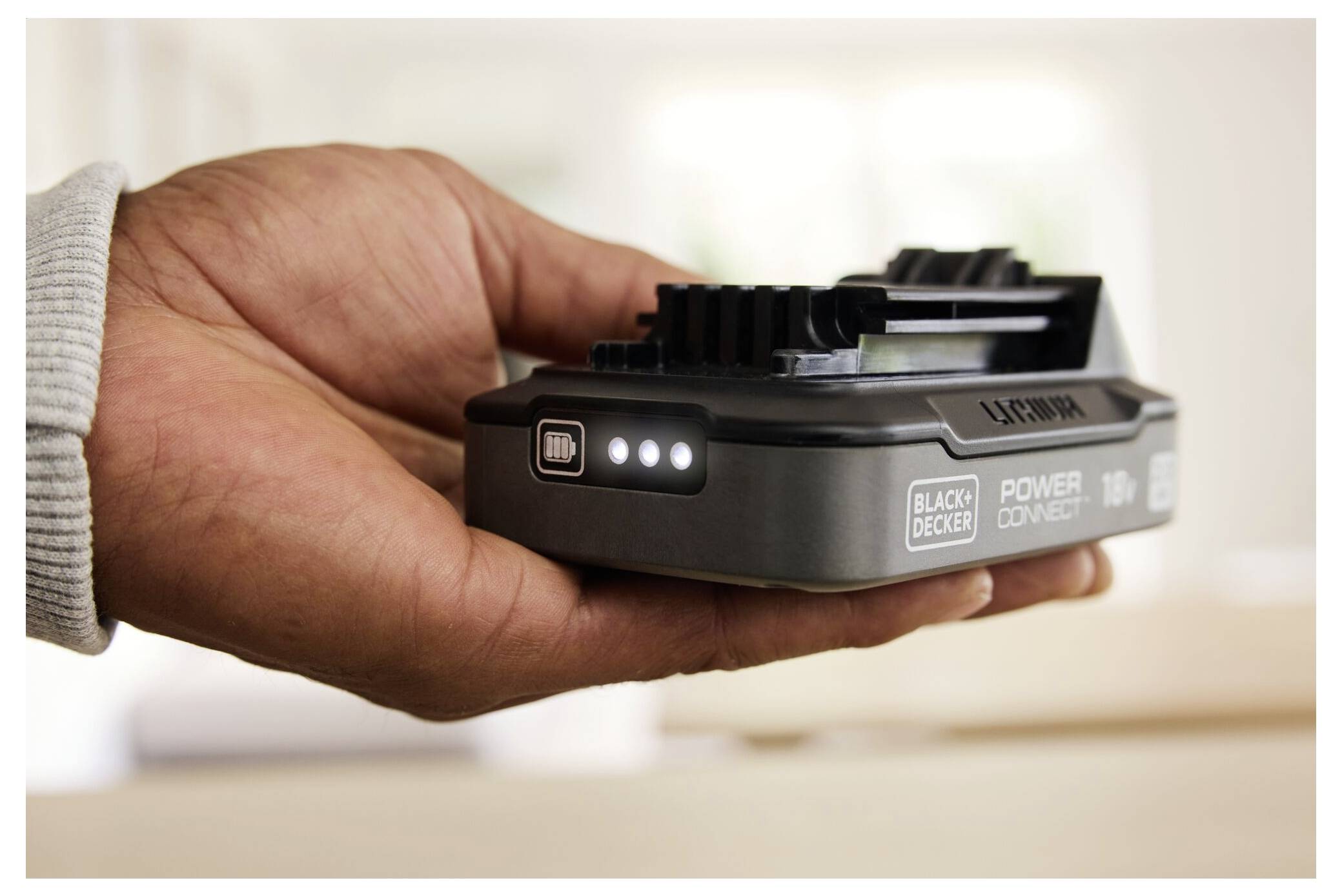 A hand holding a Black+Decker 18V battery with LED charge indicators lit, suggesting the battery is either charging or fully charged.