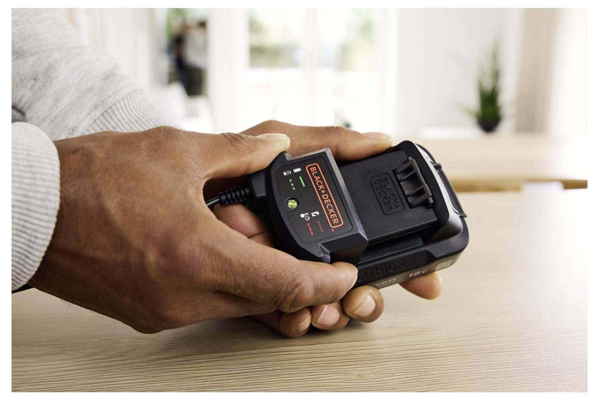 A close-up of hands holding a Black+Decker battery charger with lights indicating charging status. The charger rests on a wooden surface.