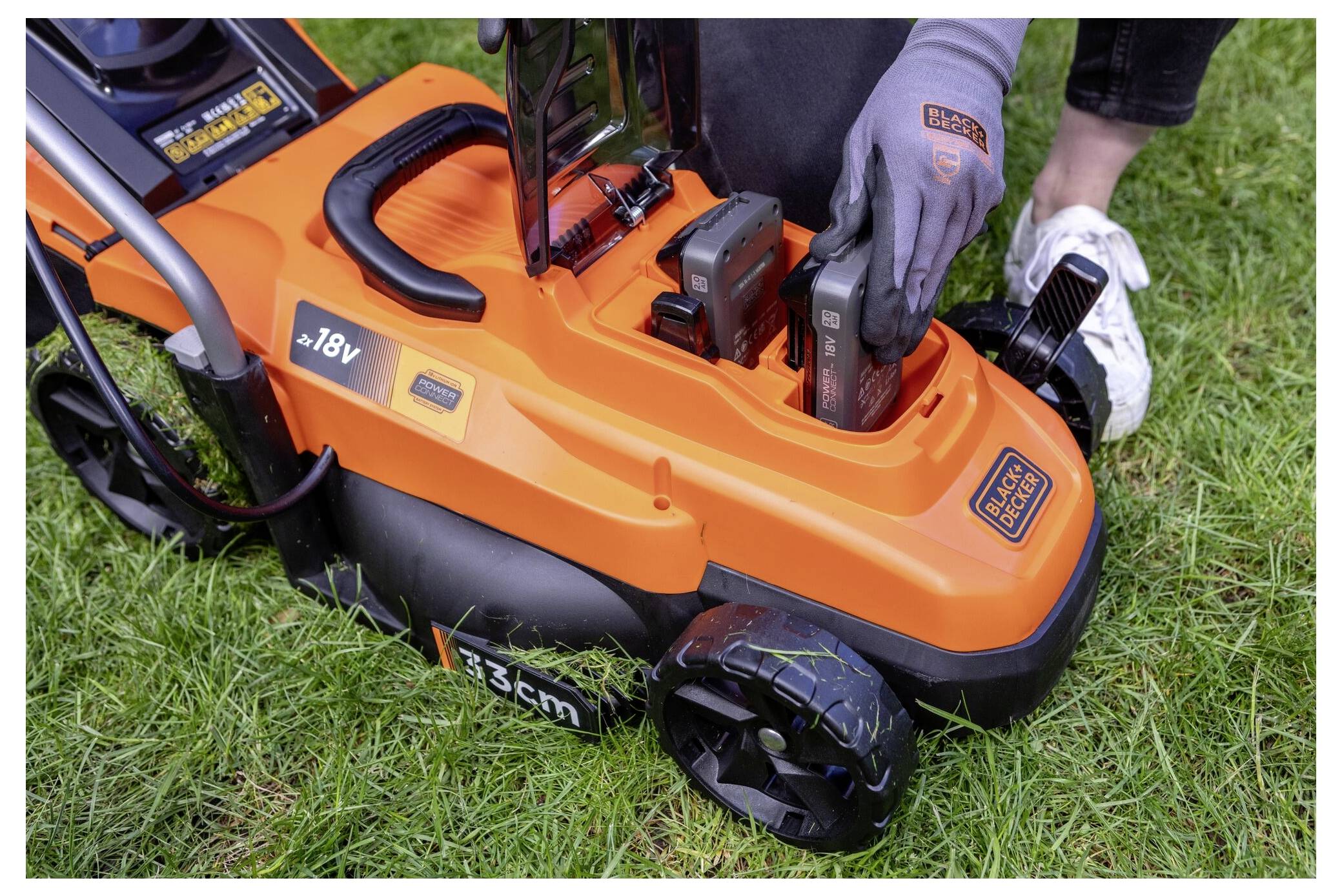 'A person is inserting a battery into an orange and black 18V lawn mower on grass. The mower has 36 cm written on the side.'