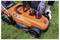 'A person is inserting a battery into an orange and black 18V lawn mower on grass. The mower has 36 cm written on the side.'