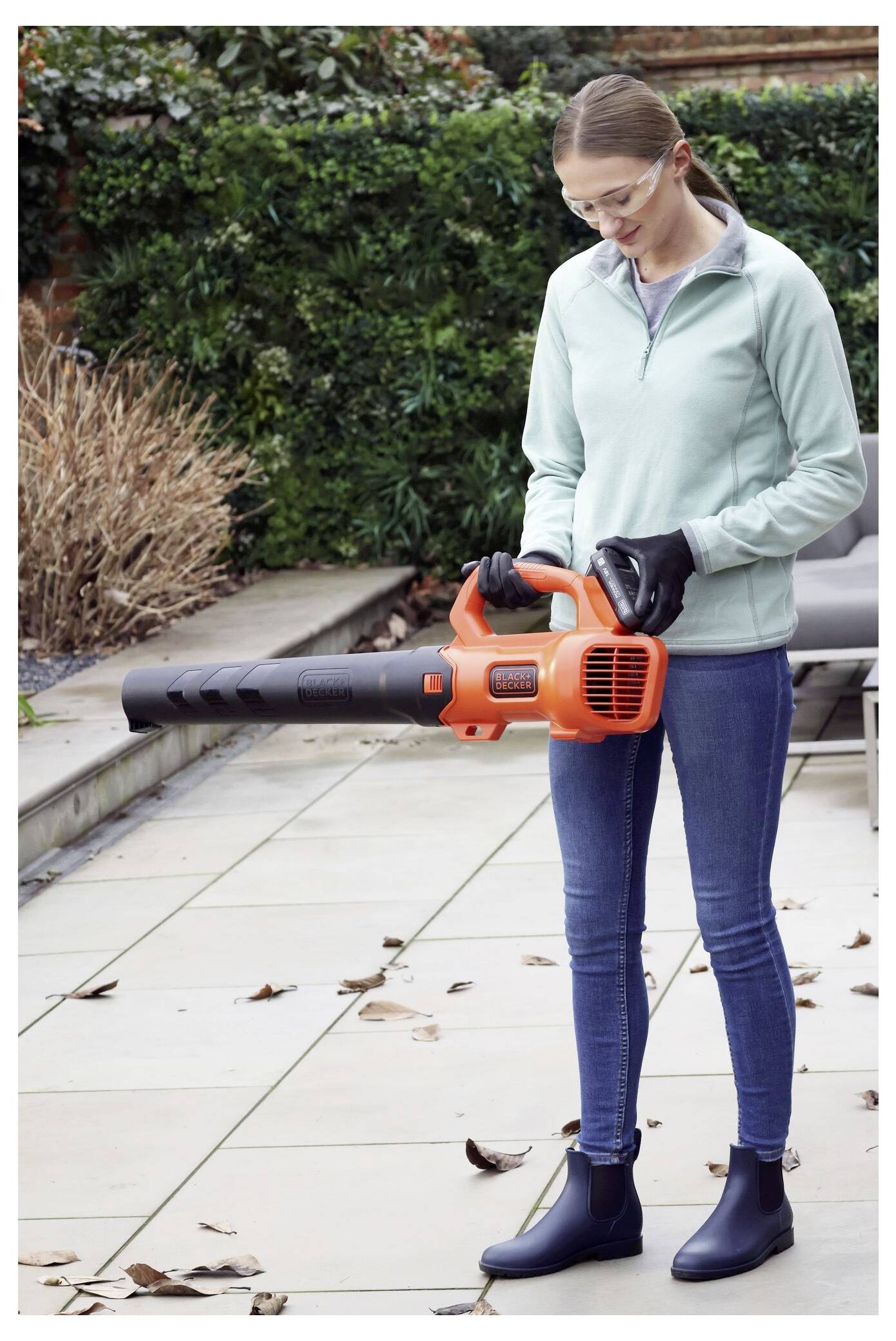 A person wearing safety goggles and gloves holds an orange leaf blower on a patio with fallen leaves.