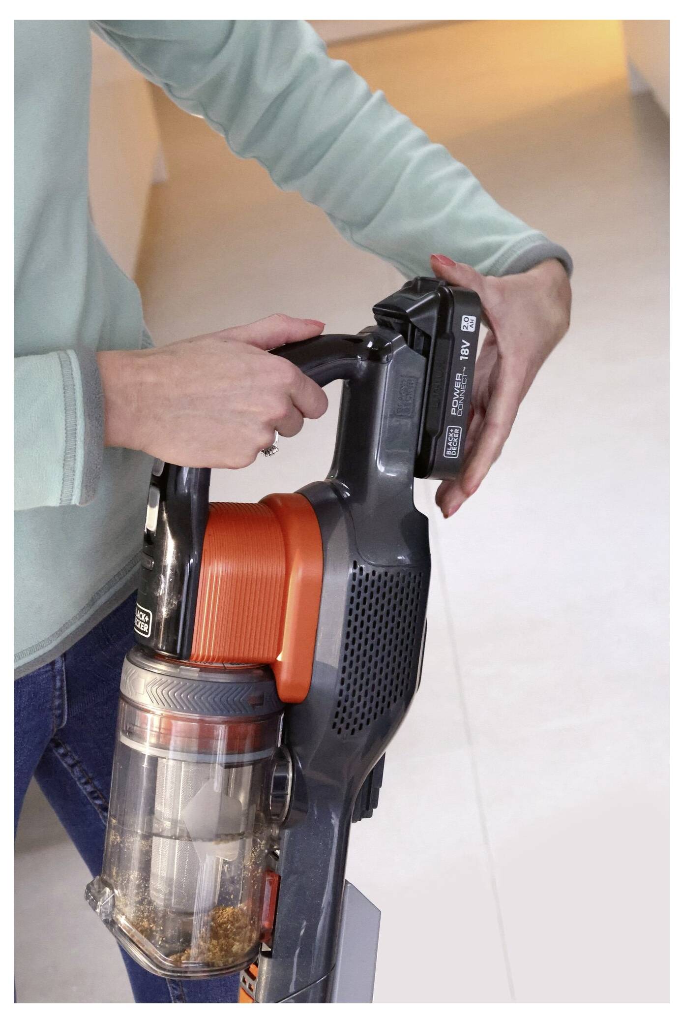A person holds an upright vacuum cleaner, demonstrating the removal or insertion of a battery pack. The focus is on the battery area.