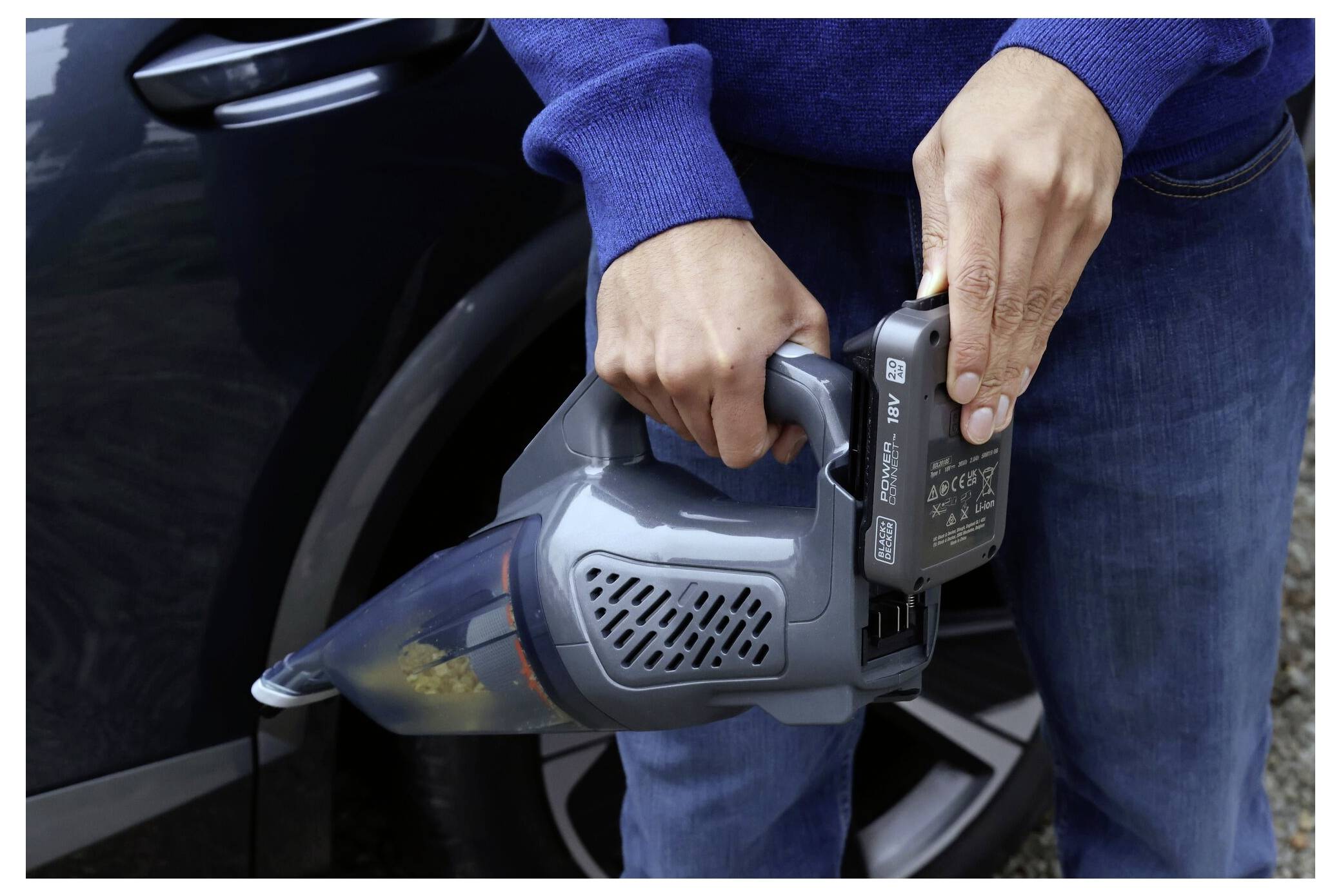 A person in a blue sweater is using a handheld vacuum cleaner to clean a car with a dark exterior.