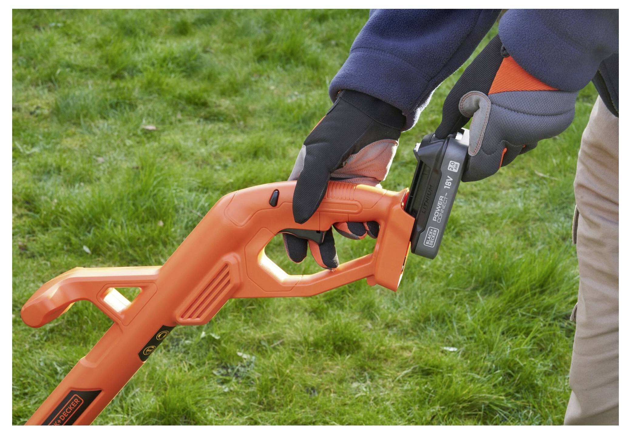 A person wearing gloves holds the handle of an orange garden tool on grass, demonstrating its battery attachment process.