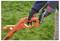 A person wearing gloves holds the handle of an orange garden tool on grass, demonstrating its battery attachment process.