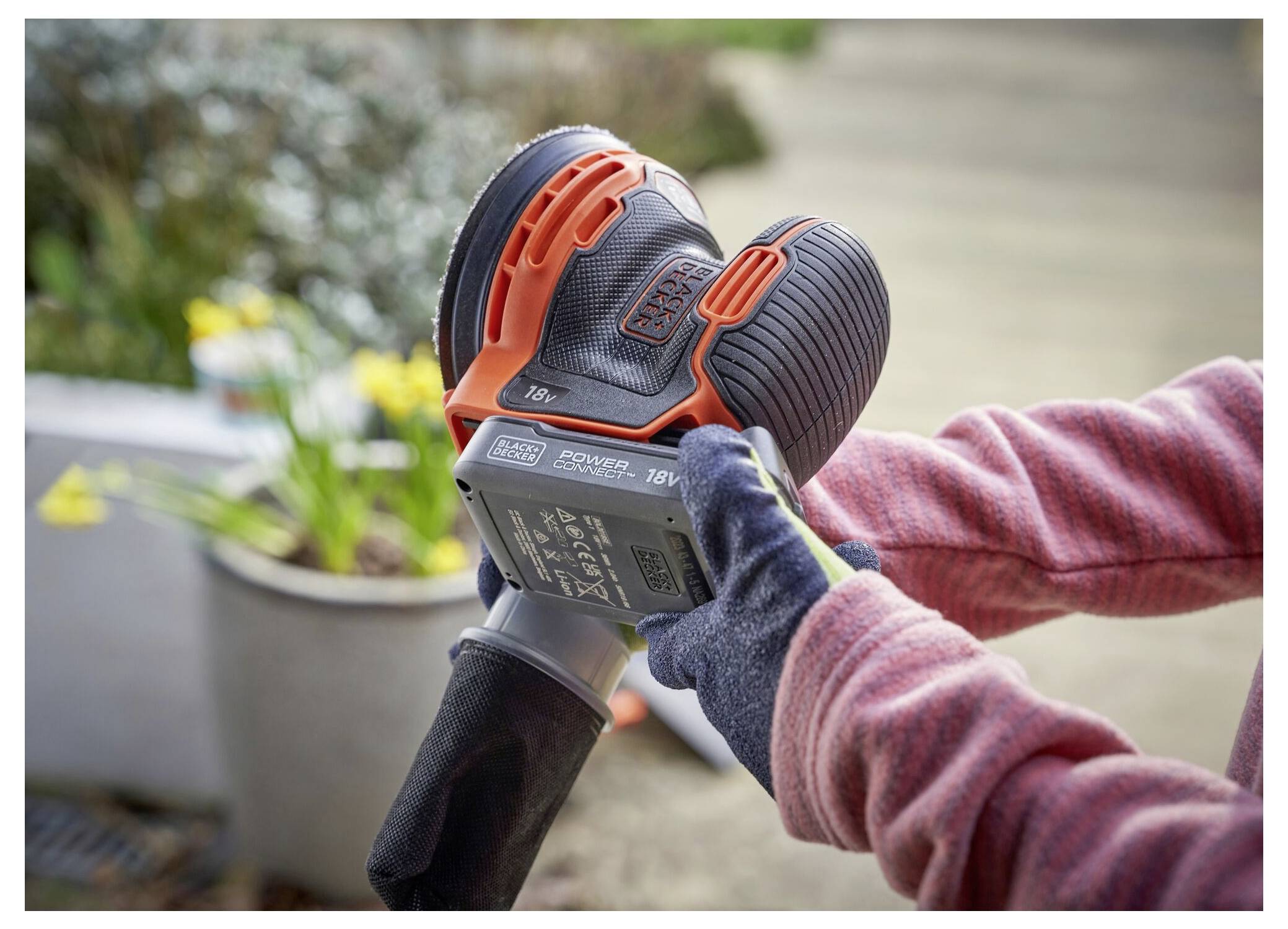 'A person wearing gloves uses a handheld electric sander on a surface, with potted flowers in the blurred background.'