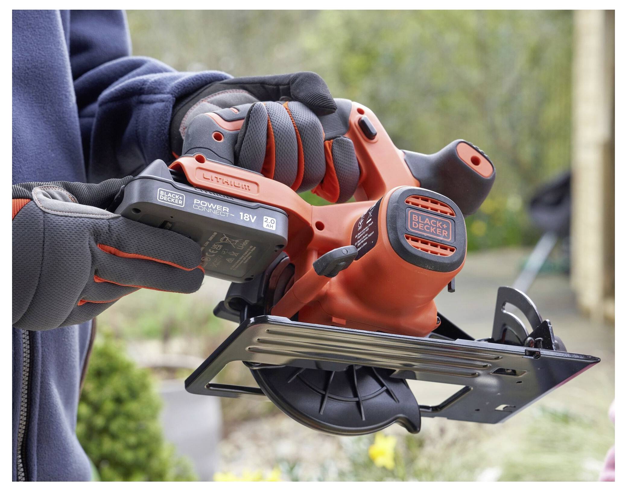 A person wearing gloves holds a cordless Black+Decker circular saw with a battery pack, standing outdoors with greenery in the background.