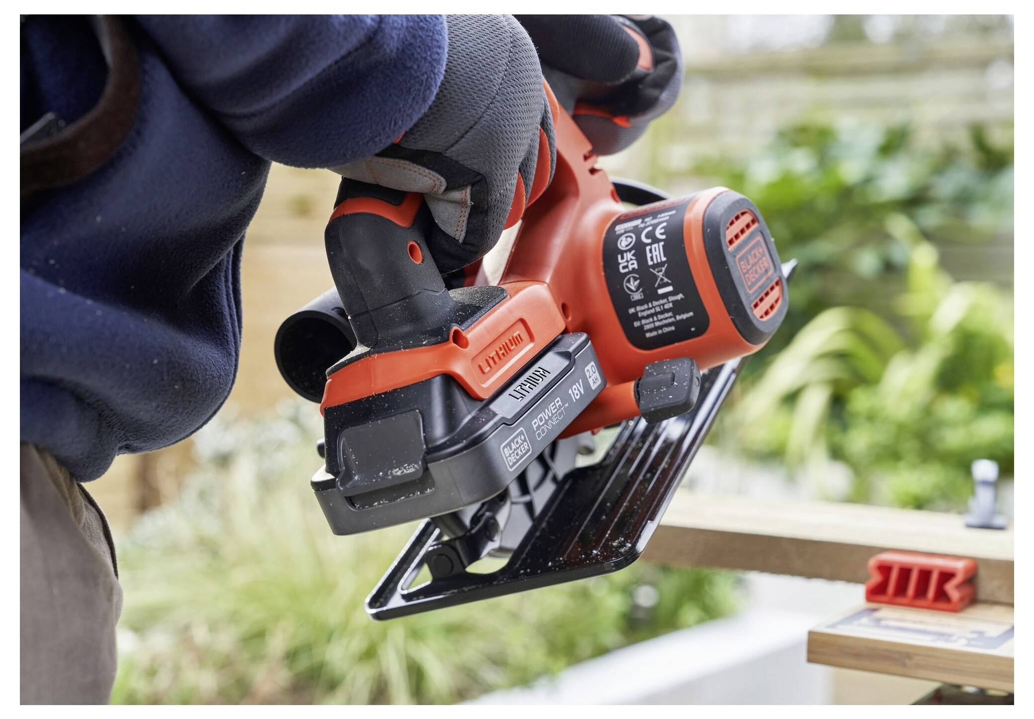 A person using a red and black power tool to cut a piece of wood outdoors, wearing gloves and a blue jacket, with greenery in the background.