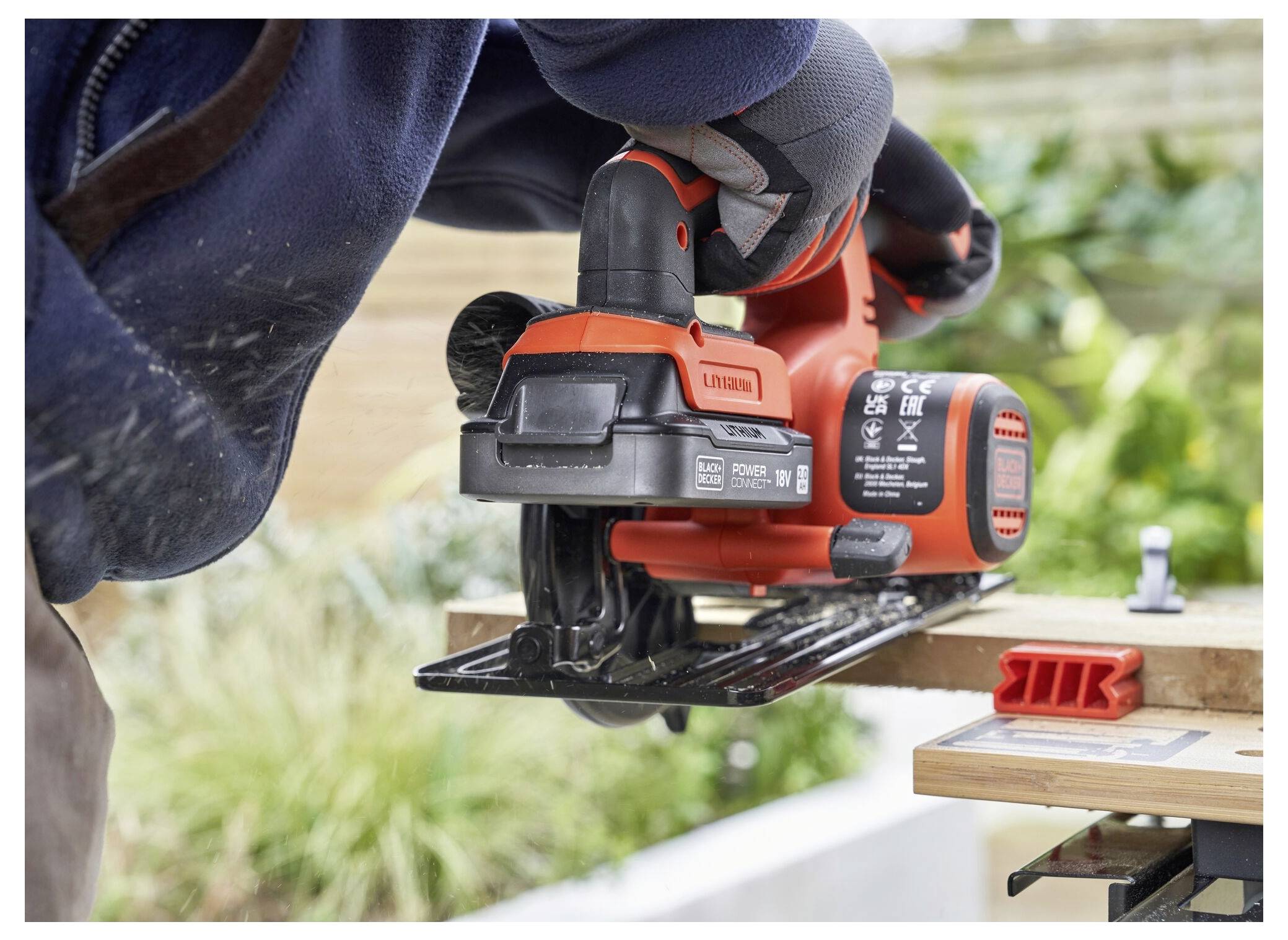 A person wearing gloves uses an orange electric jigsaw to cut a wooden board outdoors, surrounded by greenery in the background.