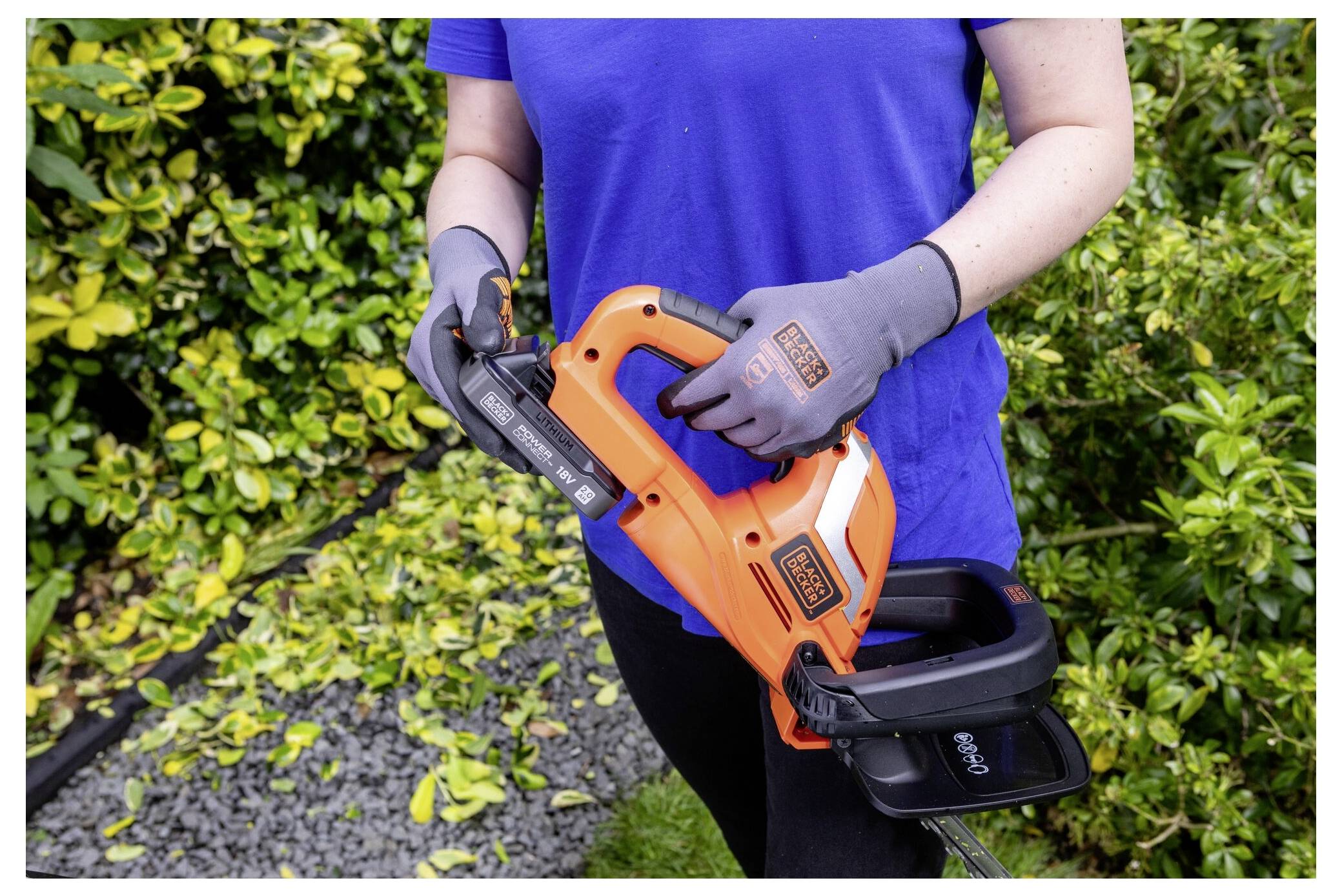 'A person operating an orange hedge trimmer in a garden, surrounded by trimmed green bushes. The individual wears gloves and a blue shirt.'