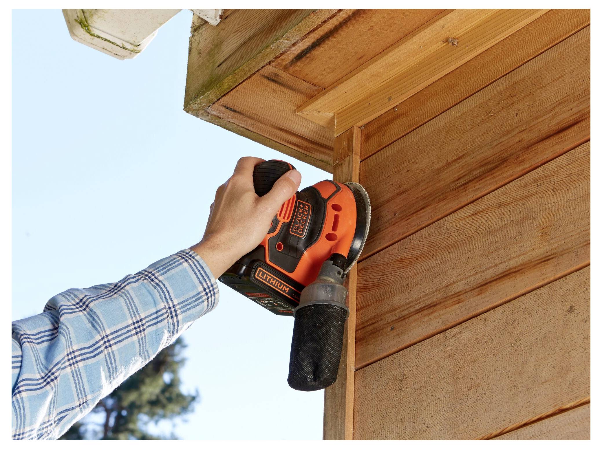 A person in a blue plaid shirt uses a hand-held electric sander on a wooden exterior corner, smoothing the surface.