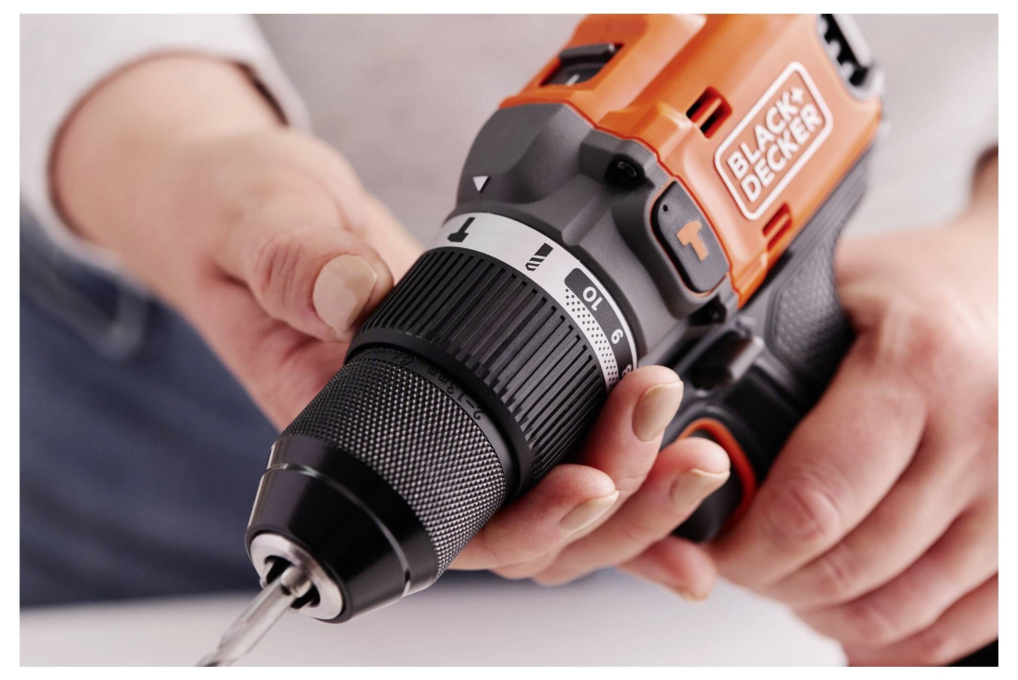 A person uses an orange cordless drill, adjusting the torque settings. The drill is branded 'BLACK+DECKER' and is held with both hands.