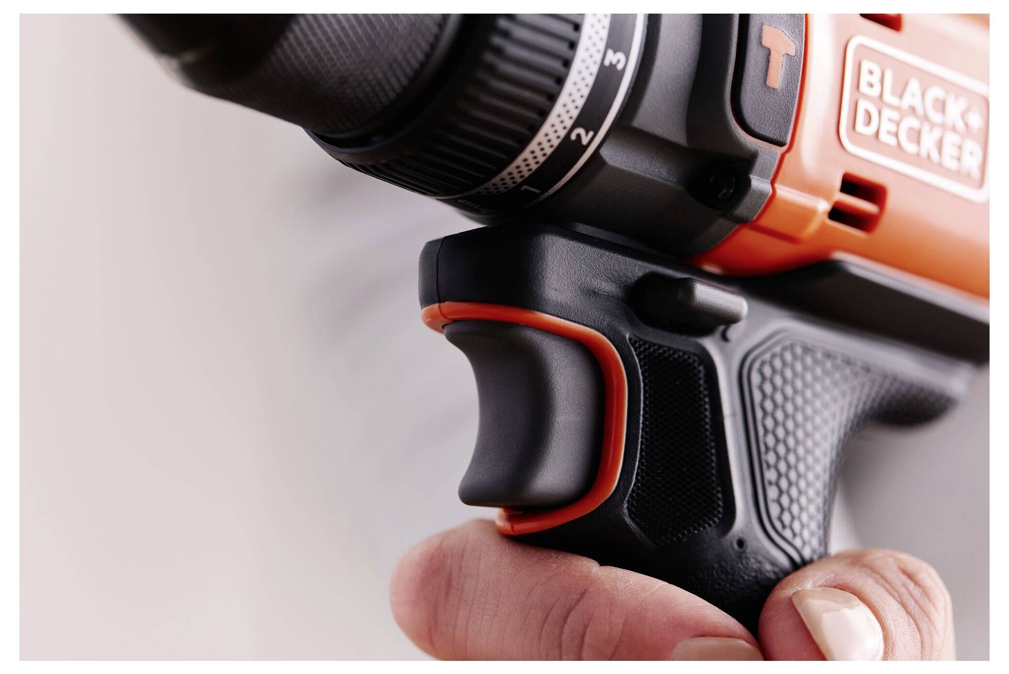 Close-up of a hand holding an orange and black drill, focusing on the grip and trigger area, with part of a logo visible.