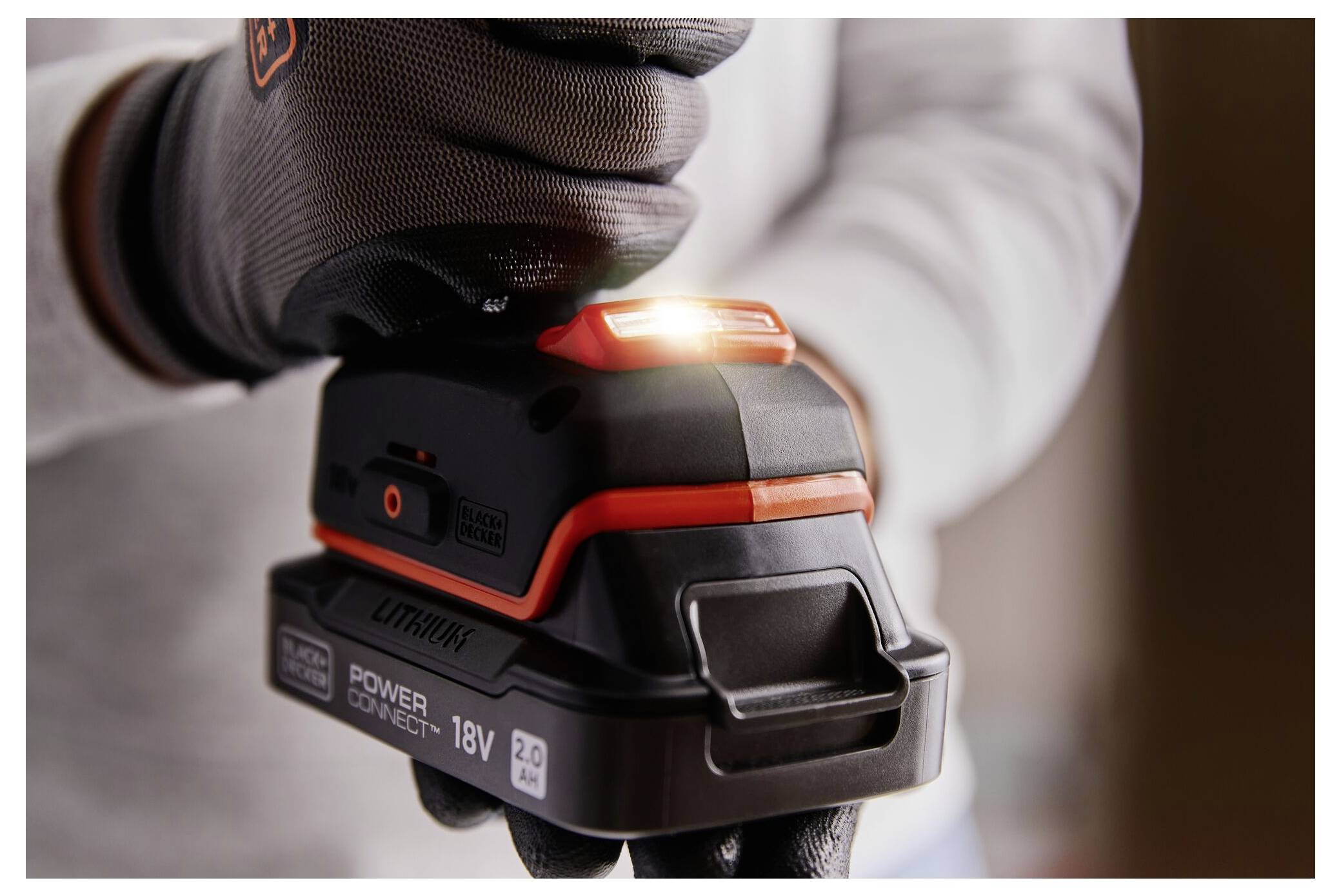 A close-up of a person adjusting an 18V lithium power tool battery, wearing gloves, with a glowing indicator light on the battery.