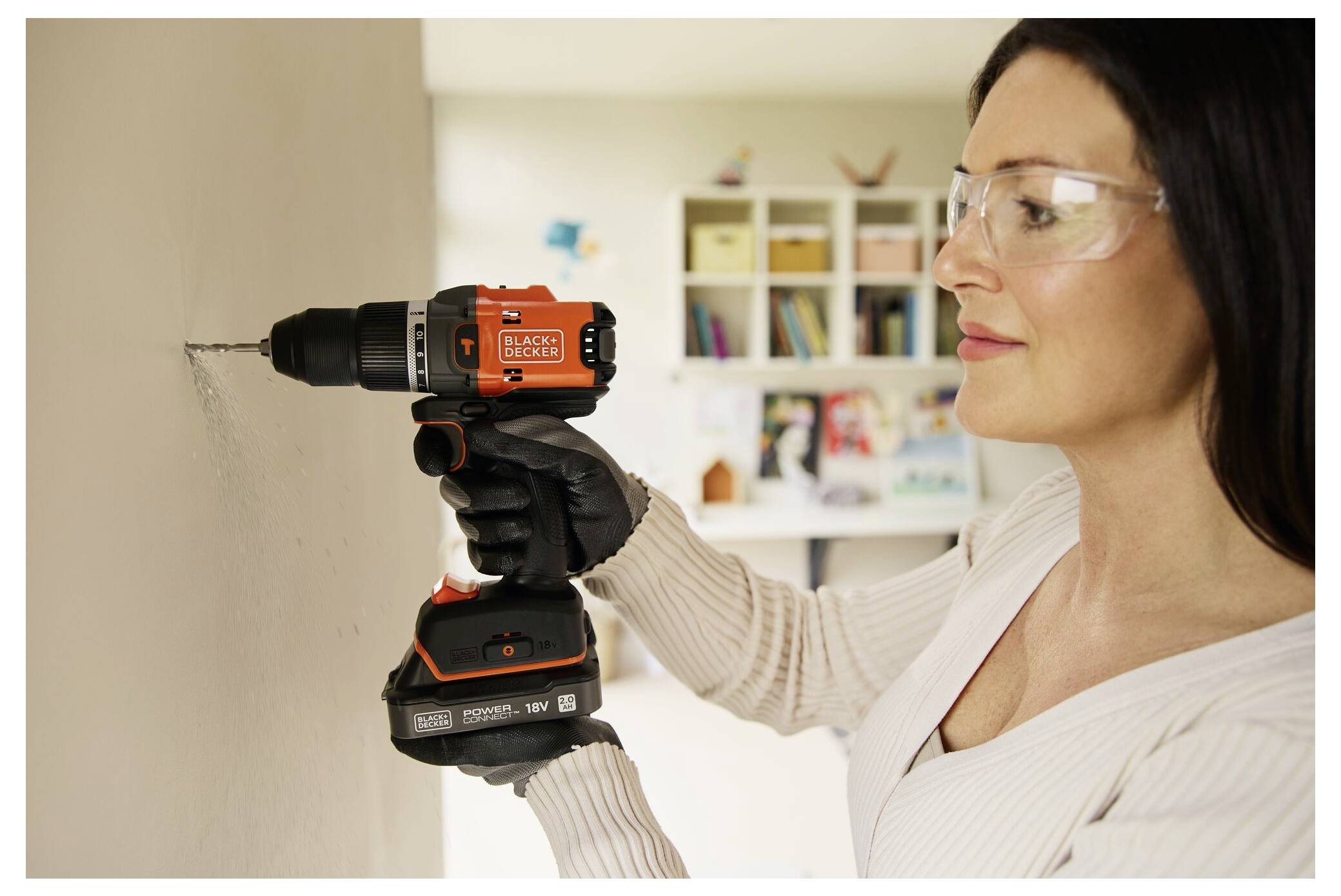 A person wearing safety goggles and gloves uses a power drill on a wall in a home setting, with shelves and books visible in the background.