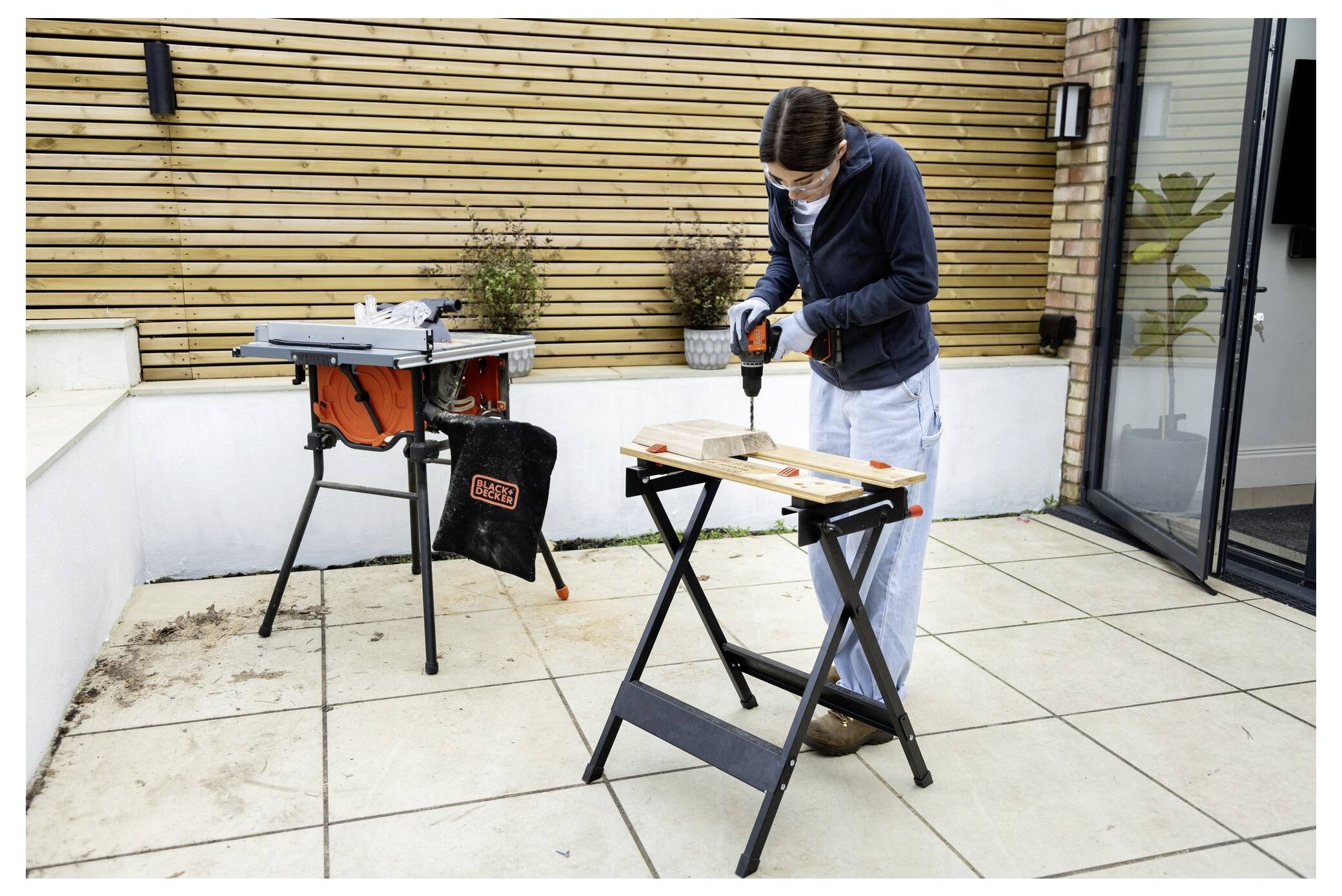 A person uses a power drill on a wooden board outdoors. A saw is set up nearby. They are in a courtyard with modern decor.