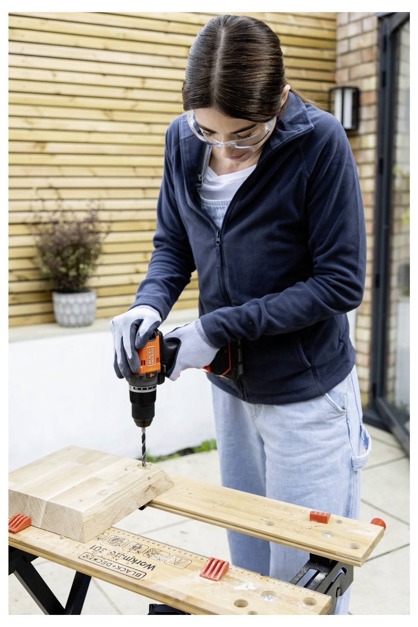 A person in a navy jacket and gloves uses a cordless drill to create a hole in a wooden block on a workbench.