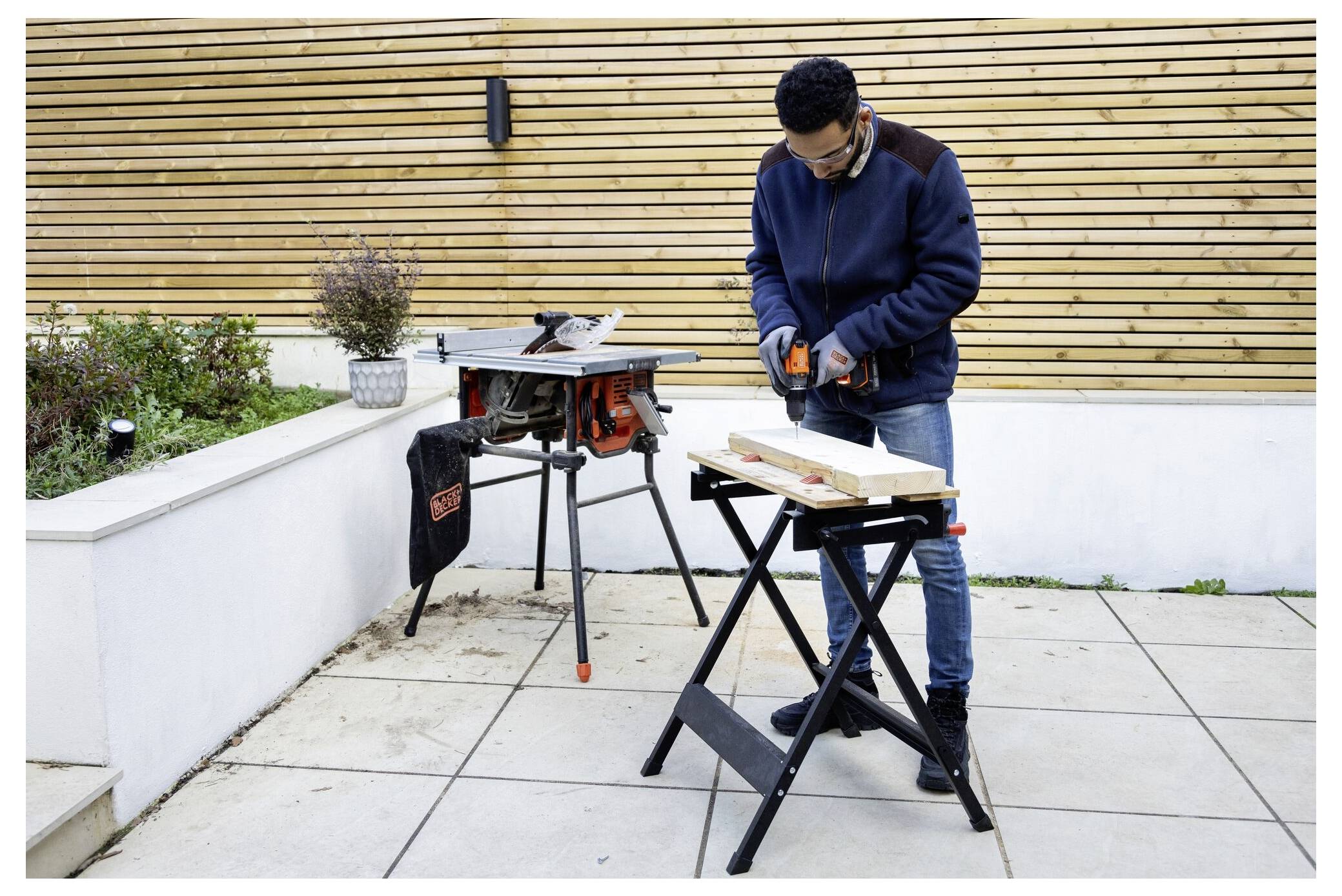 Man using a power tool on wood, standing on a patio with a table saw nearby, suggesting a woodworking project.