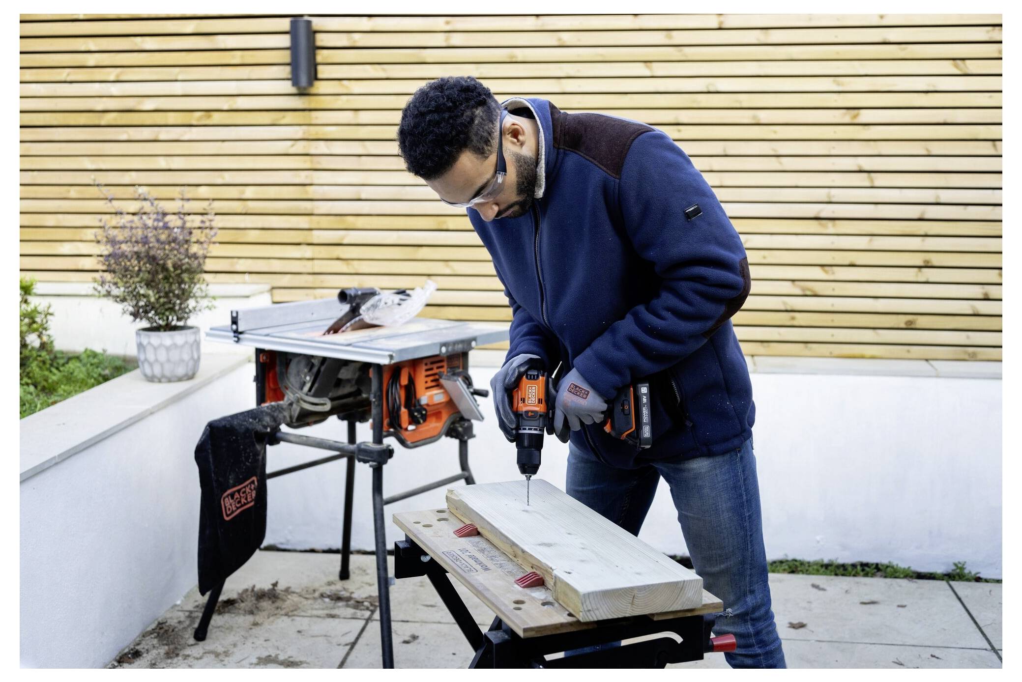 A person wearing safety glasses is drilling a hole into a wooden plank outdoors, with a power tool and workbench nearby.