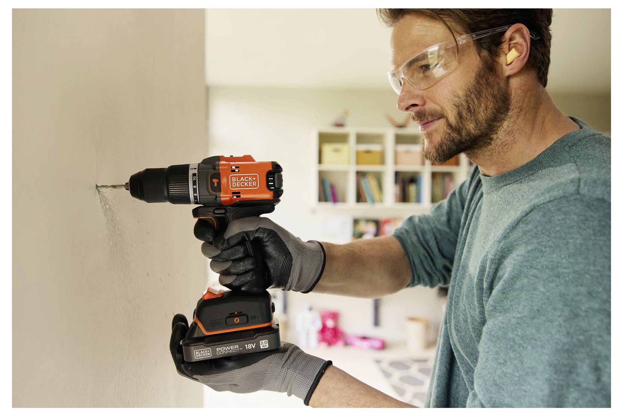A person wearing safety goggles and gloves uses a power drill to make a hole in a beige wall in a home setting, with shelves in the background.
