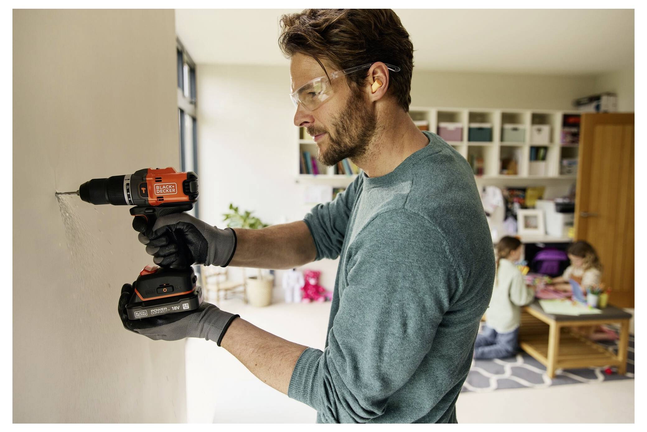A man wearing safety glasses and gloves uses a cordless drill on a wall in a living room, with children playing in the background.