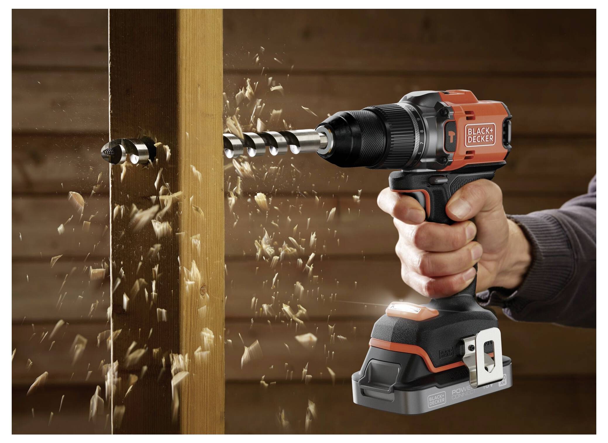 A person using a cordless Black & Decker drill to bore a hole in a wooden plank, with wood shavings flying around.