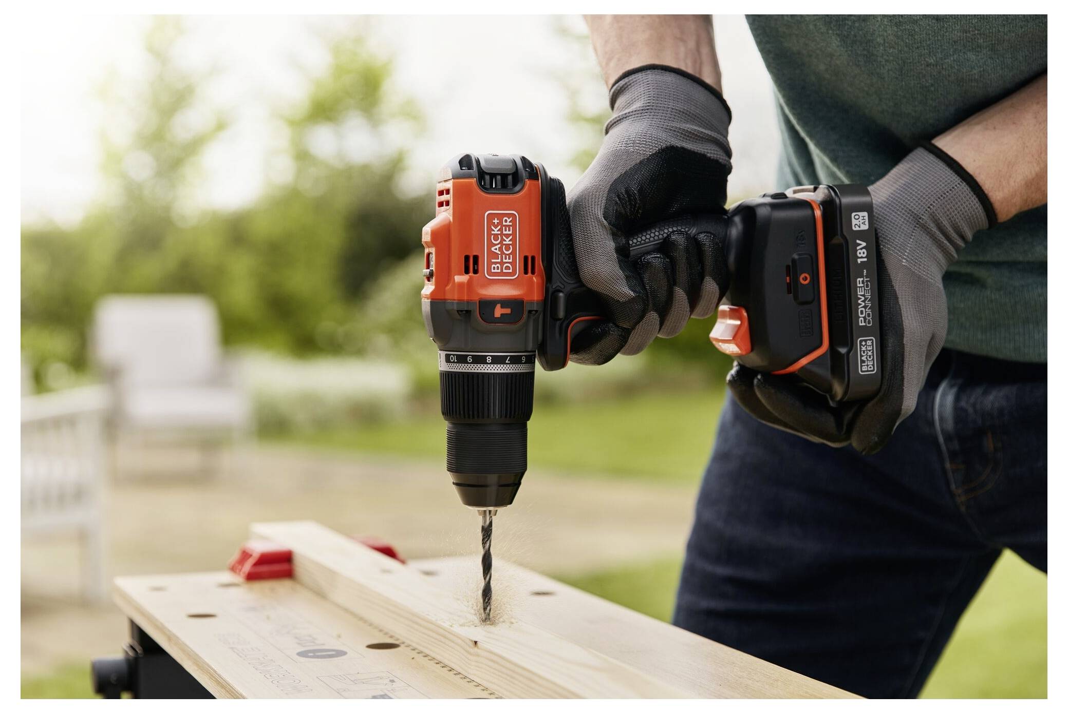 A person using an orange cordless drill to make a hole in a wooden board, wearing gloves, on a sunny day in an outdoor setting.