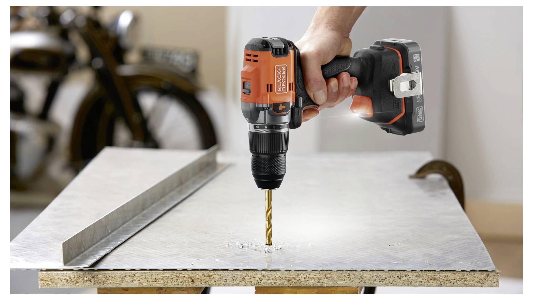 A hand operates a cordless drill with an orange casing, drilling into a metal sheet. A blurred motorcycle is visible in the background.