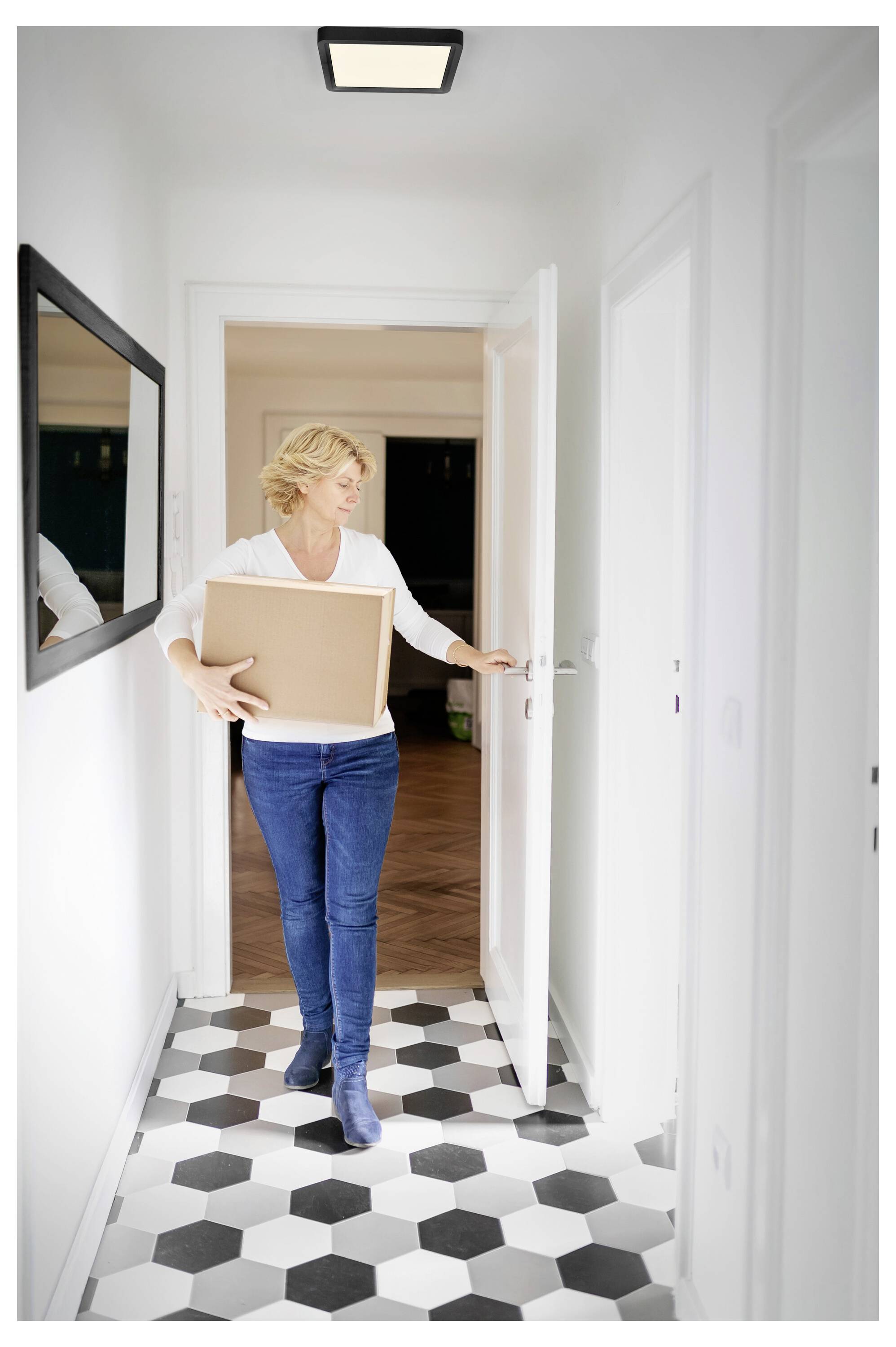 A person carrying a cardboard box walks through a brightly lit hallway with hexagonal-tiled flooring.