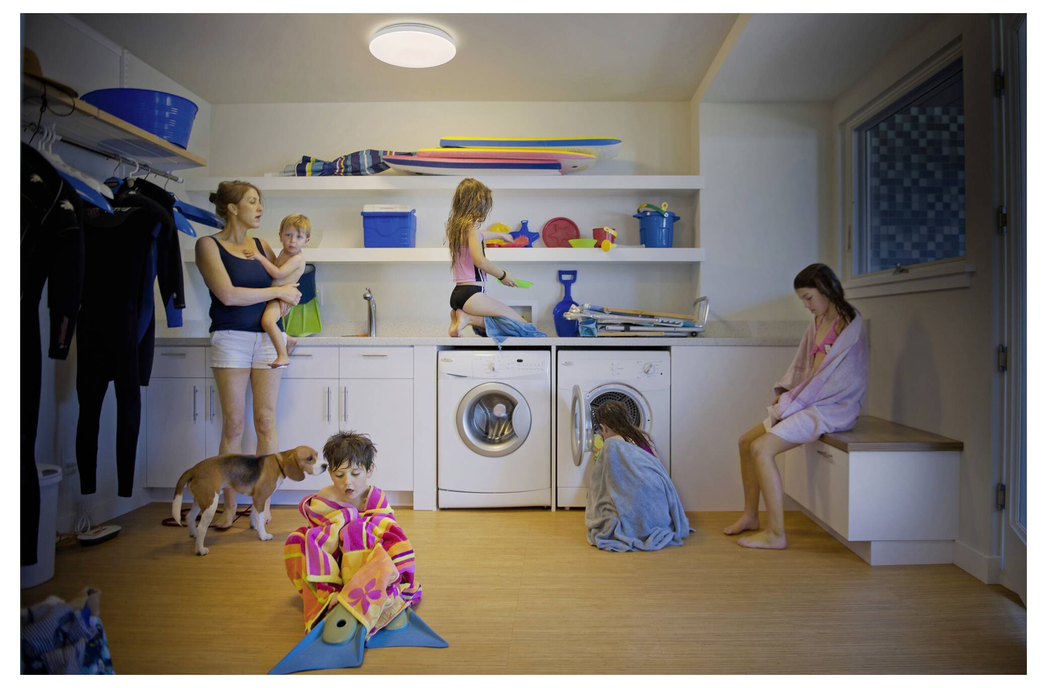 A family in a laundry room; a woman holds a child, a girl climbs a counter, a child looks inside a washing machine, and a dog is present.