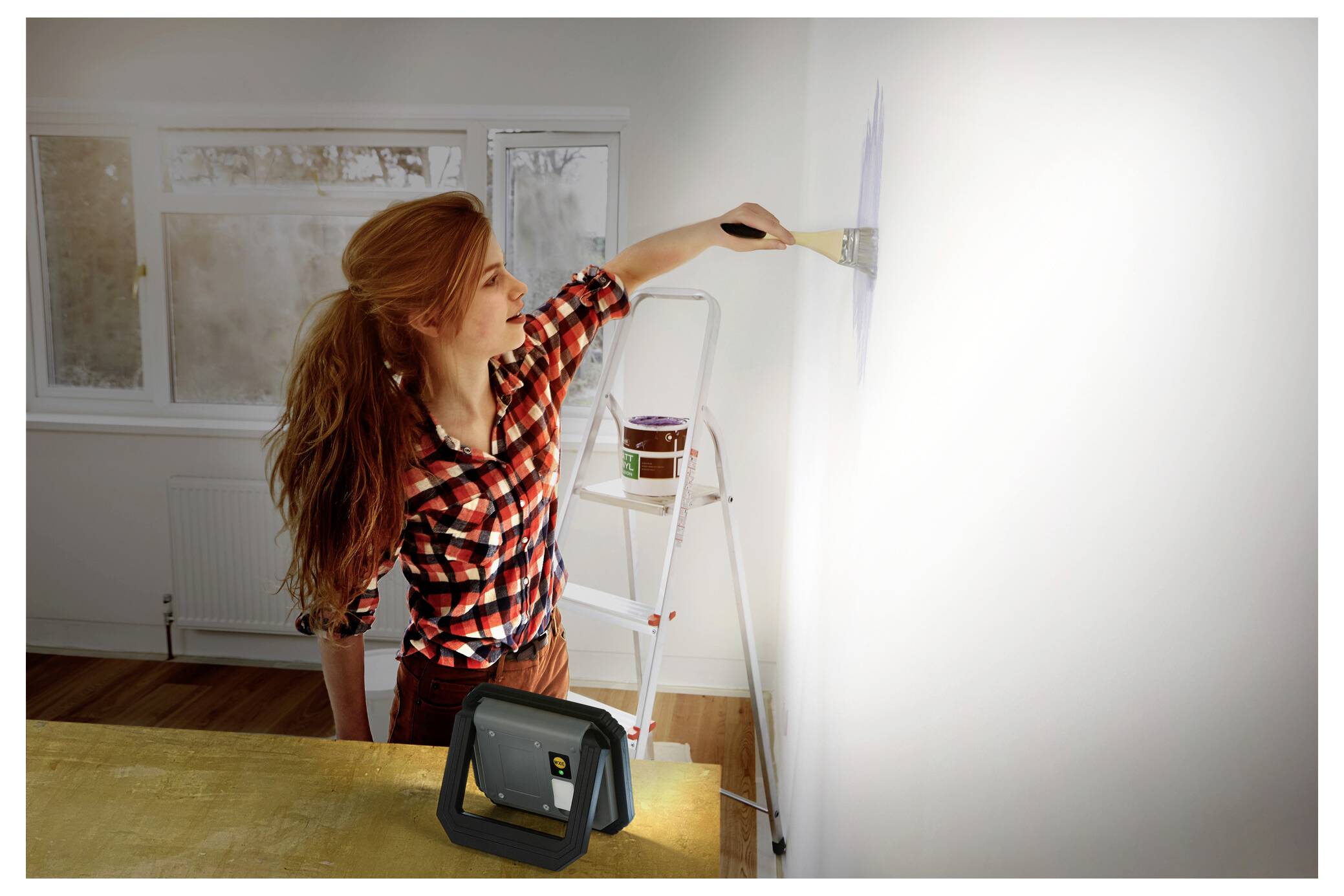 A woman with red hair, wearing a plaid shirt, is painting a white wall using a paintbrush. A ladder and paint can are nearby in the room.