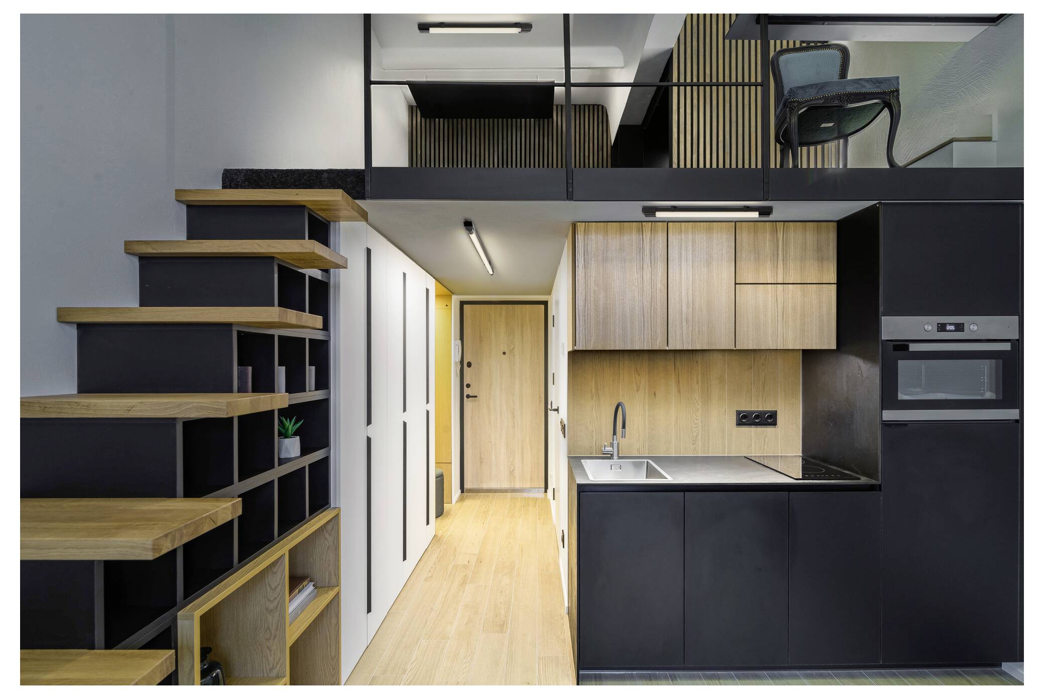 A modern loft apartment with a wooden staircase leads to an upper loft area. The main level features a compact kitchen with black and wood cabinetry.