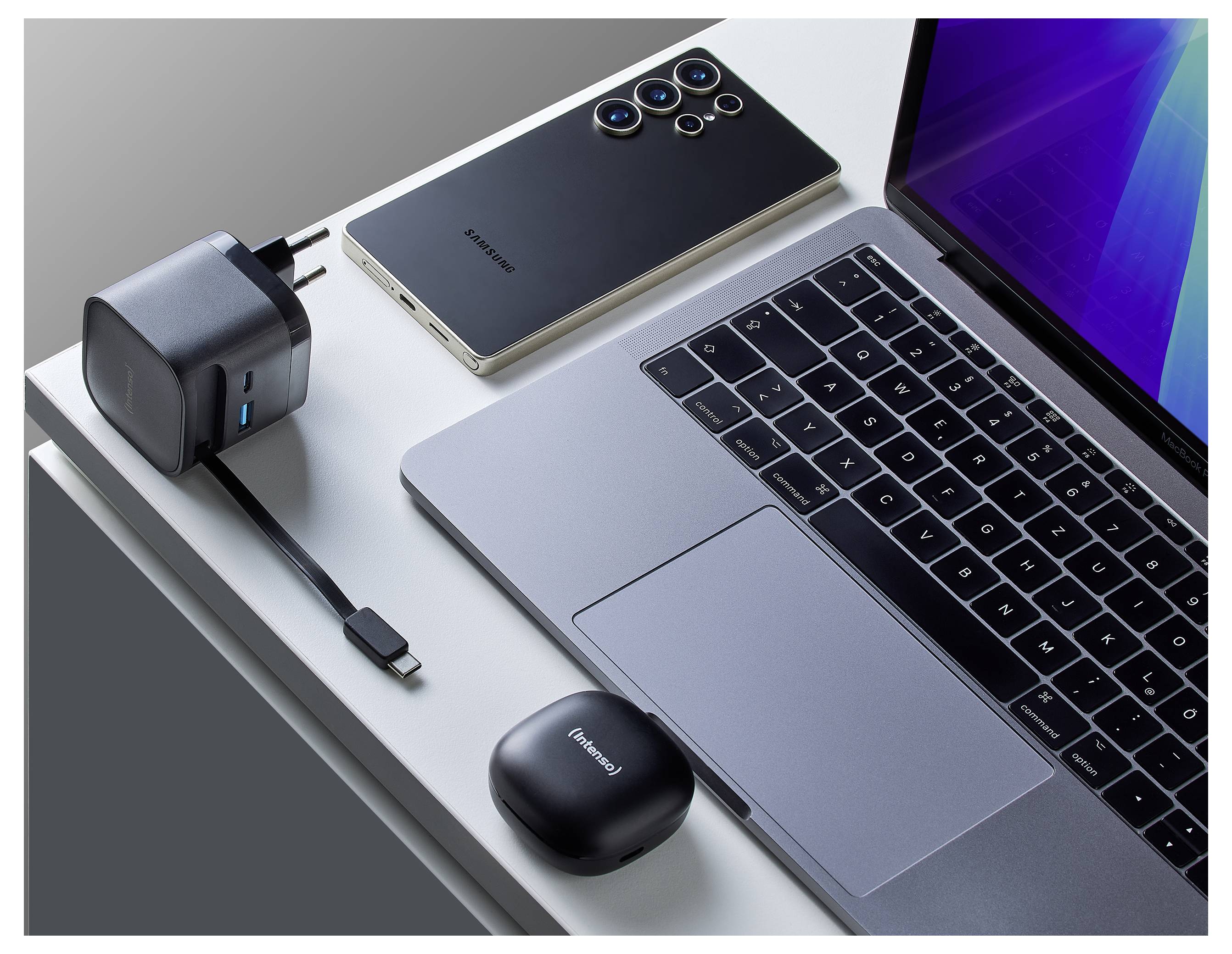 A laptop on a desk with a smartphone, a charger with a USB cable, and wireless earbuds.