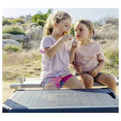 Two young girls sit on a bench outdoors sharing a snack in a sunny rocky landscape.