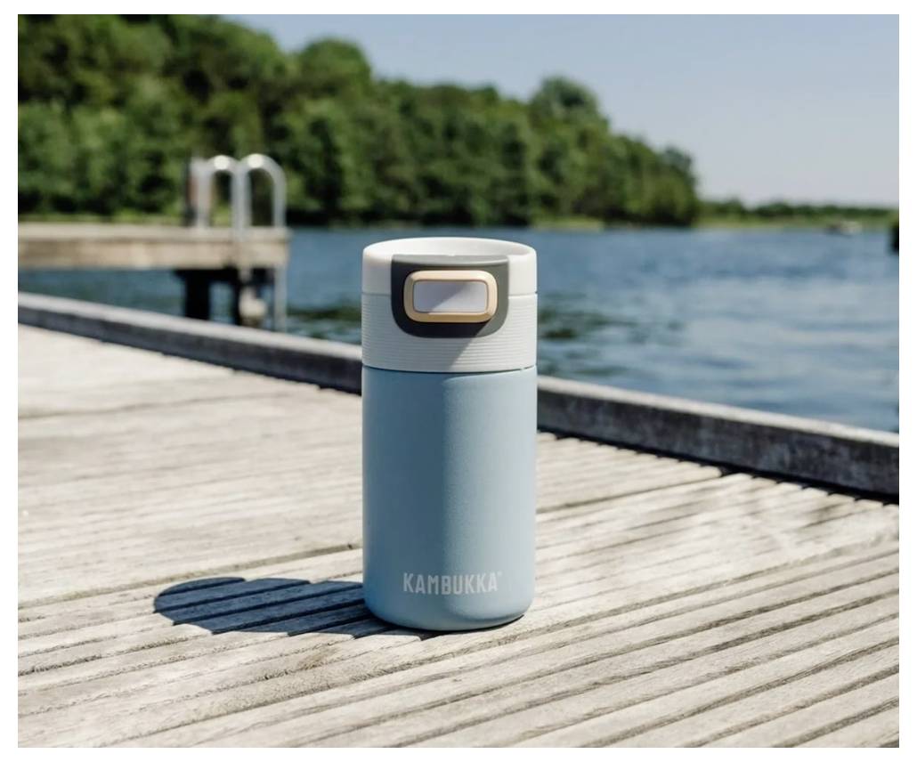 'A light blue Kambukka thermos stands on a wooden dock by a lake, surrounded by trees under a clear sky.'