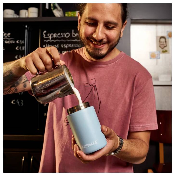 A person in a pink shirt pours milk from a metal jug into a blue 'Kambukka' cup. They're smiling, standing in a coffee shop setting.
