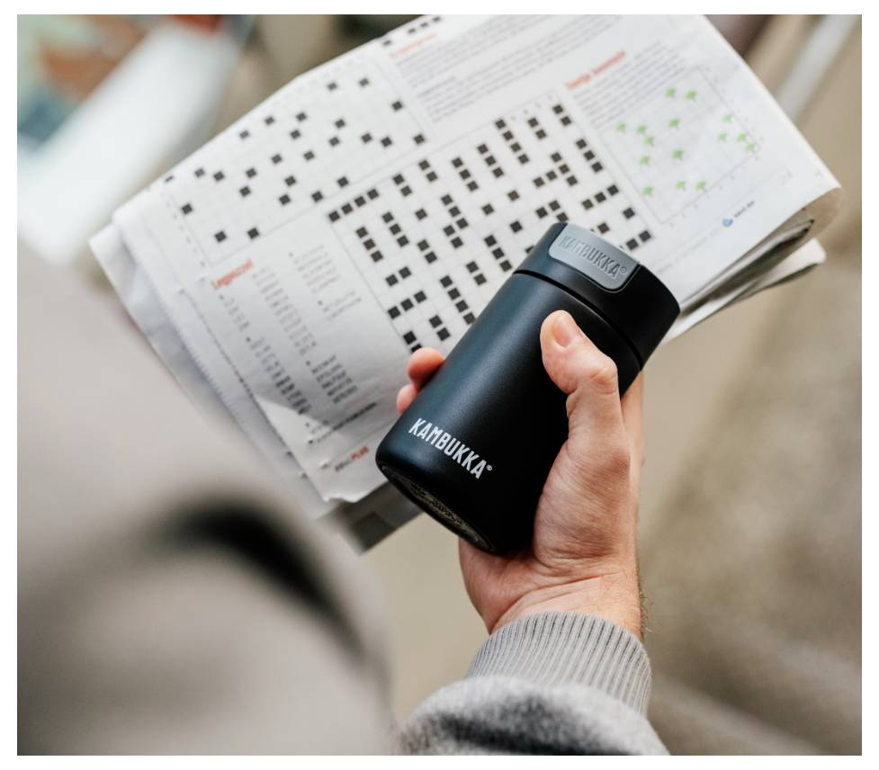 A person holds a travel mug while reading a newspaper with Sudoku puzzles and a dot plot graph, emphasizing leisure and relaxation.