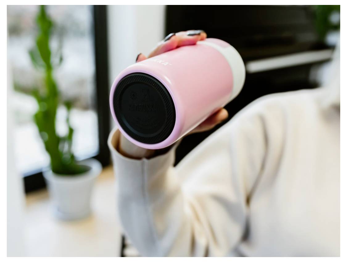 A person holds a pink portable speaker in their hand indoors, with a potted plant and a window in the background.