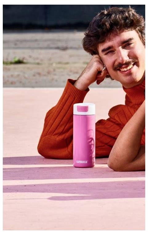 A person in an orange sweater smiles while leaning on a pink surface. In front of them is a pink water bottle with a white lid.