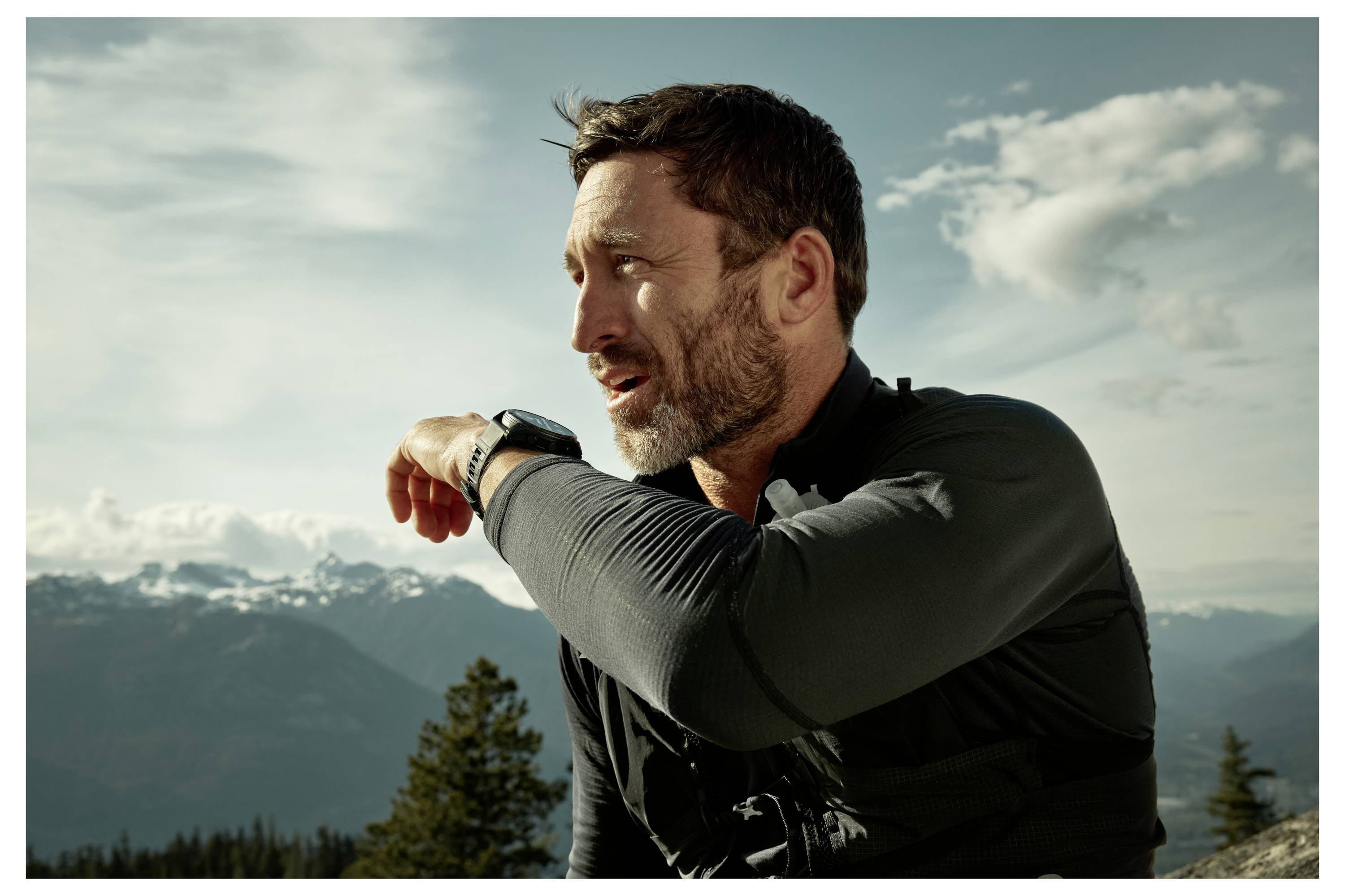 Man in outdoor setting looking at a smartwatch, with mountains and trees in the background.