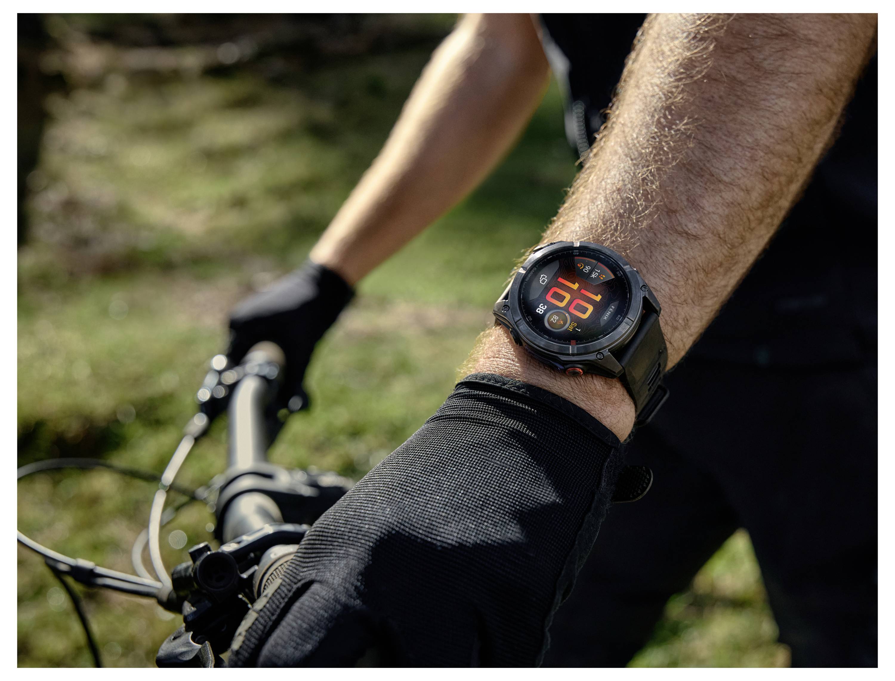 A person wearing black gloves cycles outdoors, displaying a watch with a digital screen on their wrist, indicating time and weather.