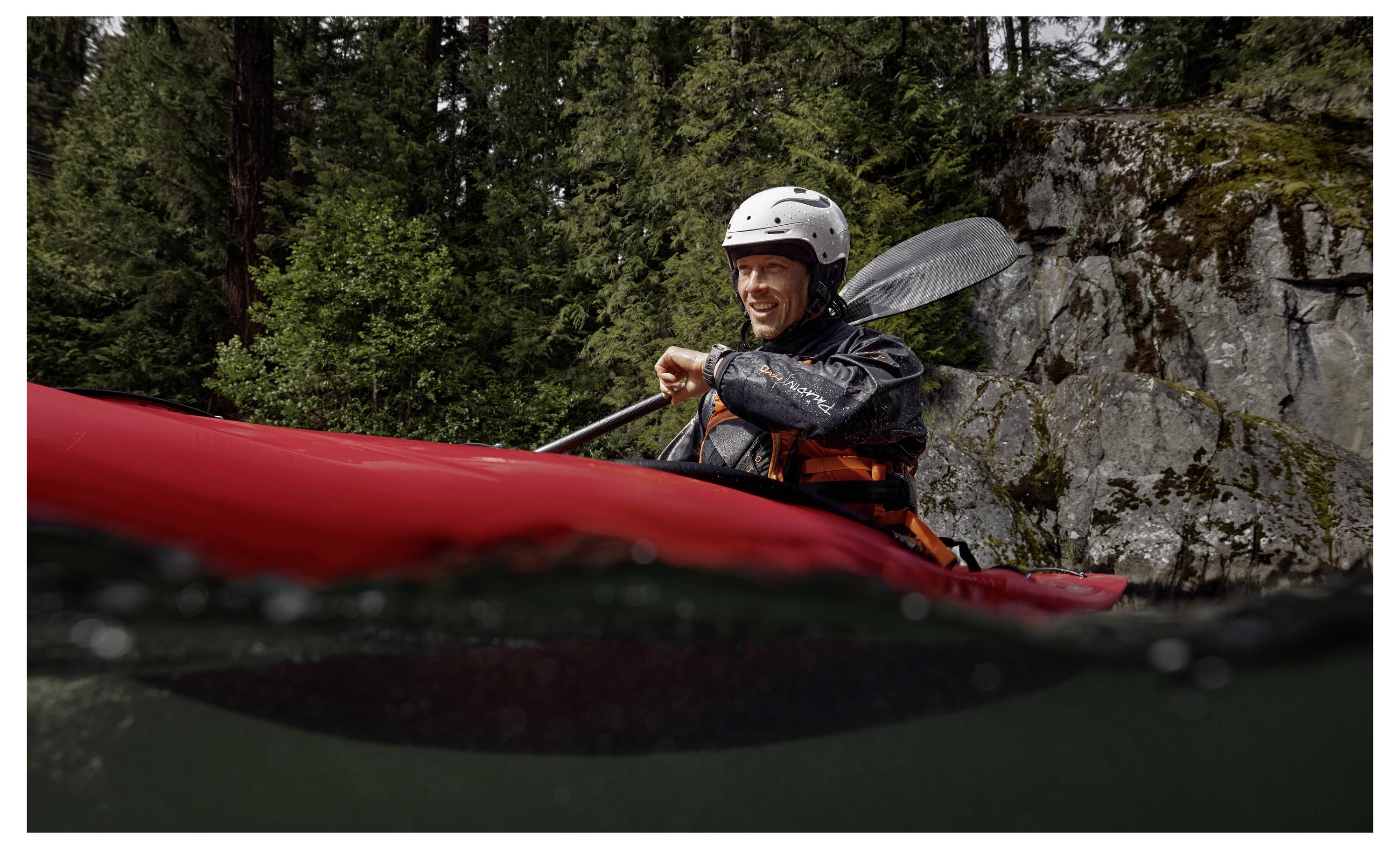 A person wearing a helmet and life jacket paddles a kayak on a river, surrounded by forest and rocky terrain.