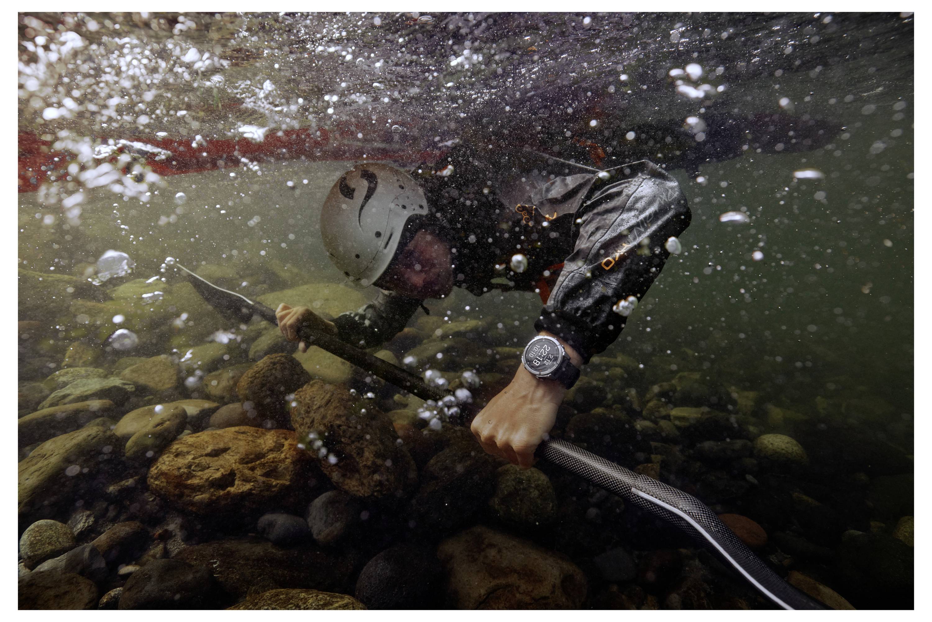 A kayaker in a helmet and life jacket is submerged underwater, holding a paddle near a rocky riverbed, with bubbles surrounding them.