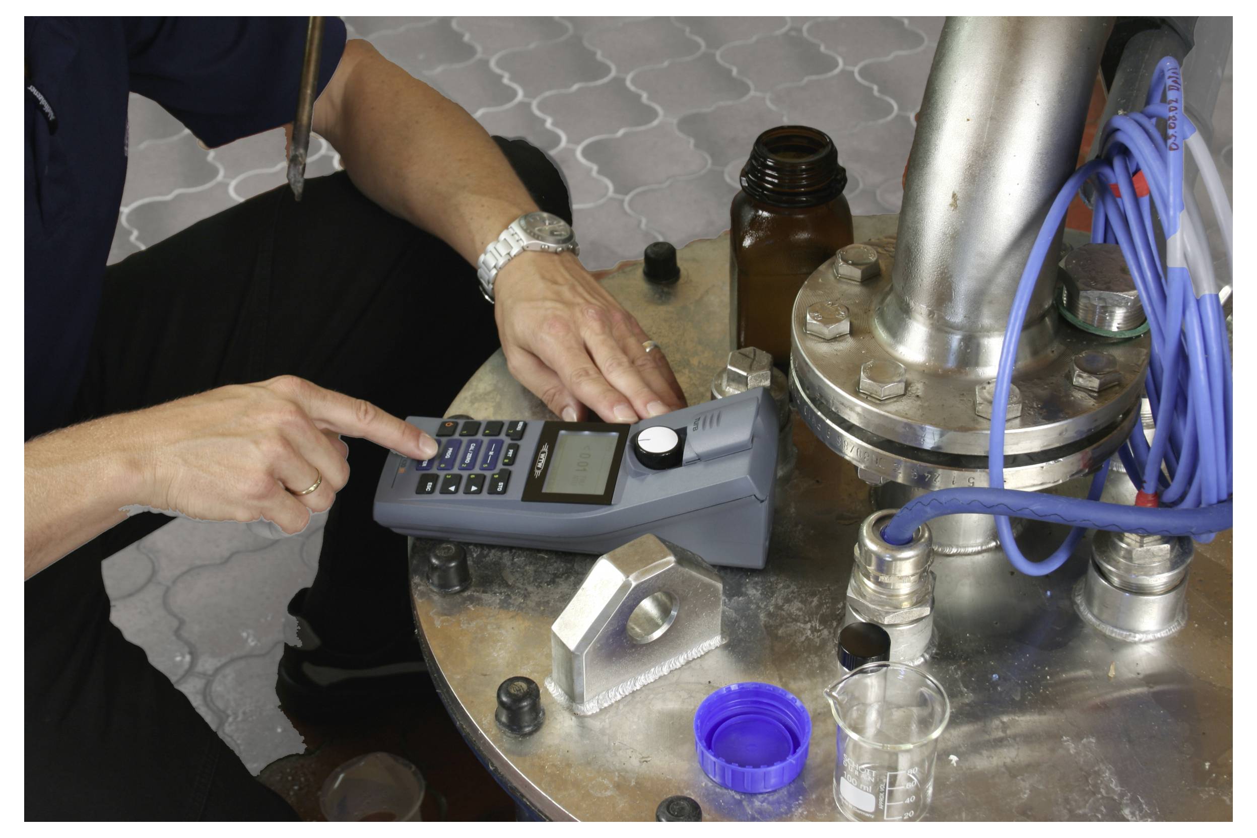 A person uses a handheld digital device to measure or monitor liquid near industrial equipment, with blue hoses and a brown bottle nearby.