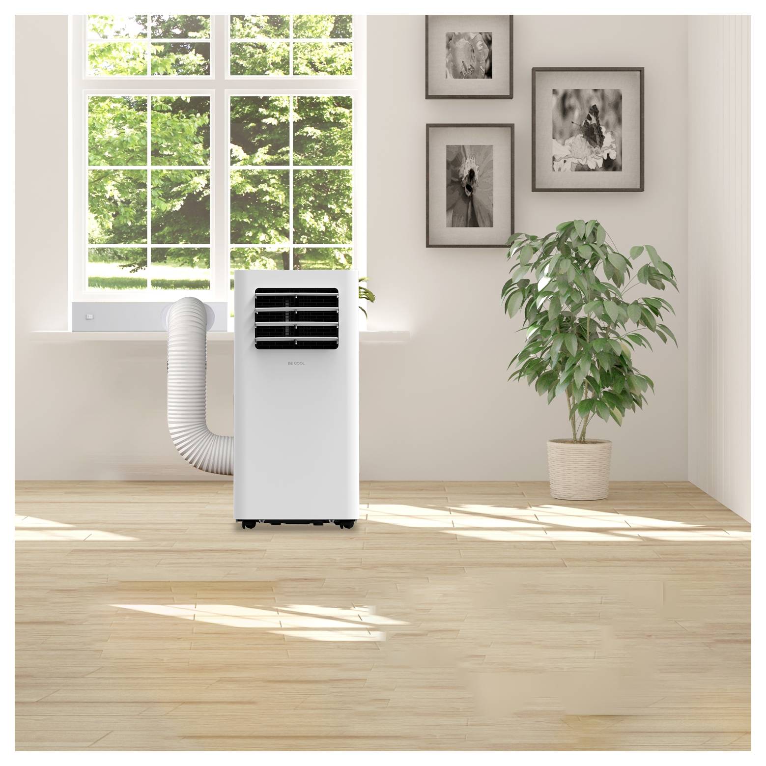A portable air conditioner stands on a wooden floor in a bright room with framed photos on the wall, a large window, and a potted plant nearby.
