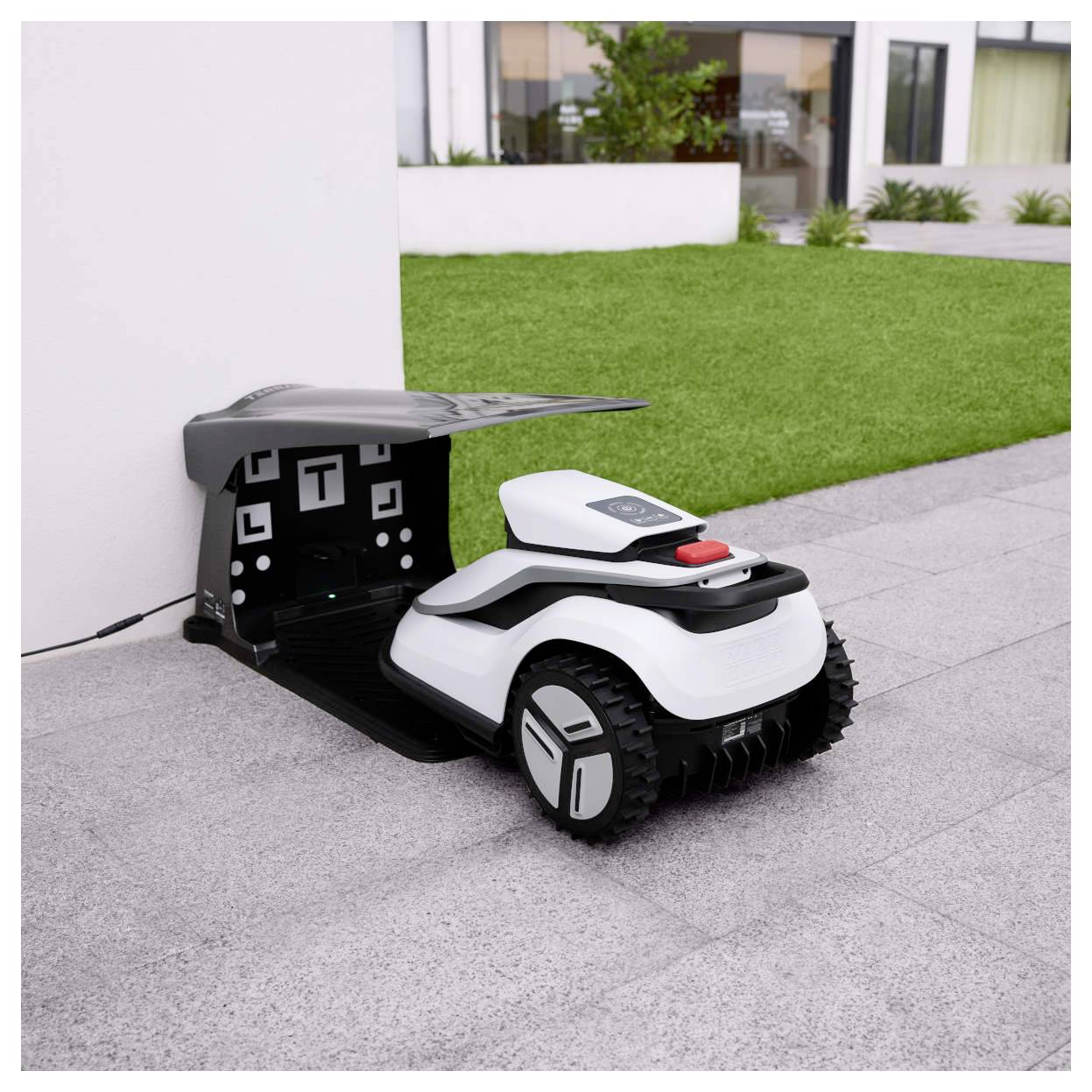 A robotic lawn mower is parked on a concrete patio near a patch of green grass, with a small shelter structure on its charging base.