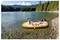 A man and woman relax in a colorful inflatable boat on a clear lake surrounded by forest, with a snow-capped mountain in the background.