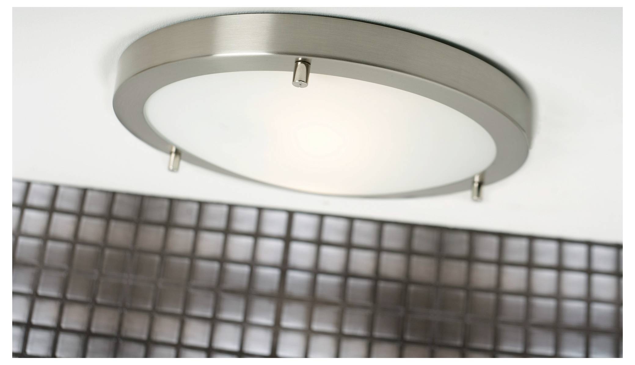 A modern ceiling light with a circular, brushed metal fixture and frosted glass, mounted above a grid of small square tiles.