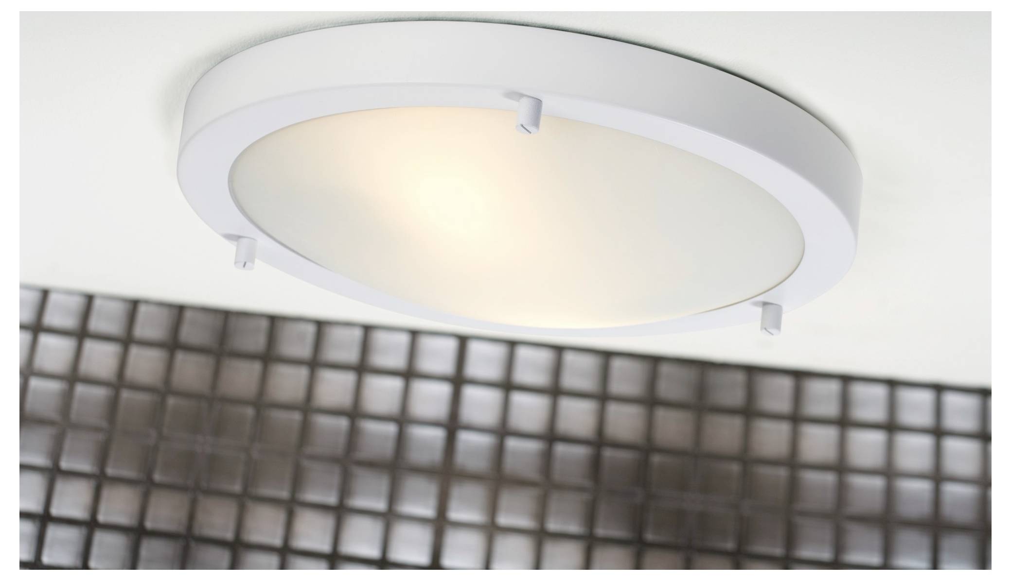 A round ceiling light fixture emits a soft glow. Below it, there is a wall with a grid pattern of reflective, square tiles.
