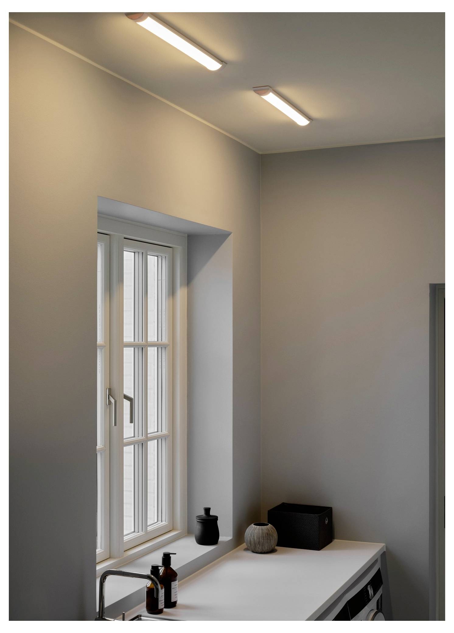 A minimalist kitchen corner with a sink under a window. Two linear ceiling lights illuminate the space. Decorative items sit on the counter.